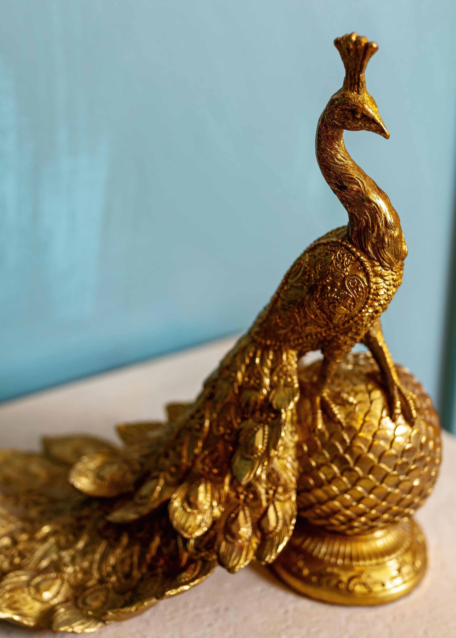 Gold peacock figurine with ornate tail, standing on a decorative base against a blue background