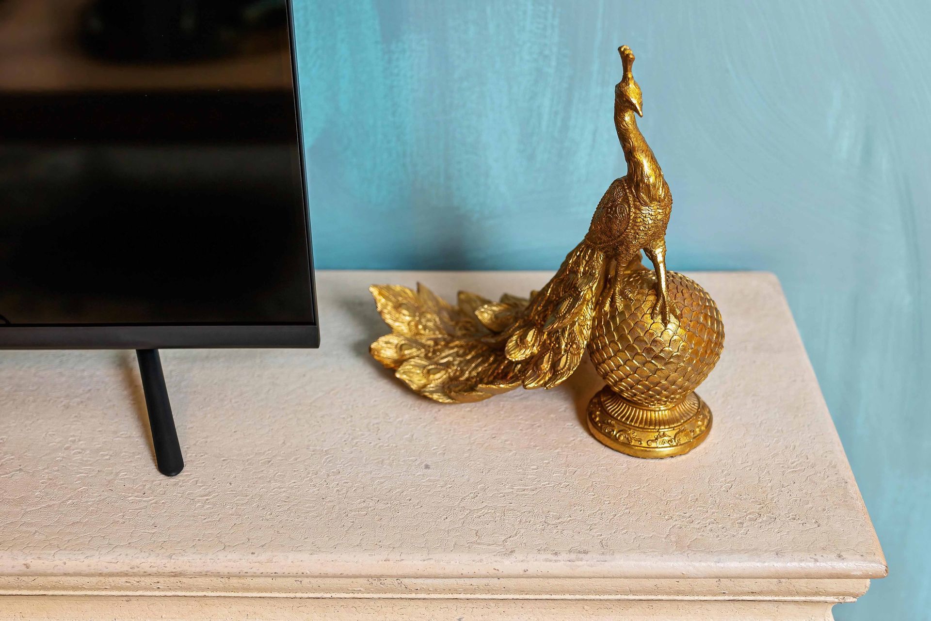 Gold peacock figurine on a beige mantel beside a TV, against a blue wall
