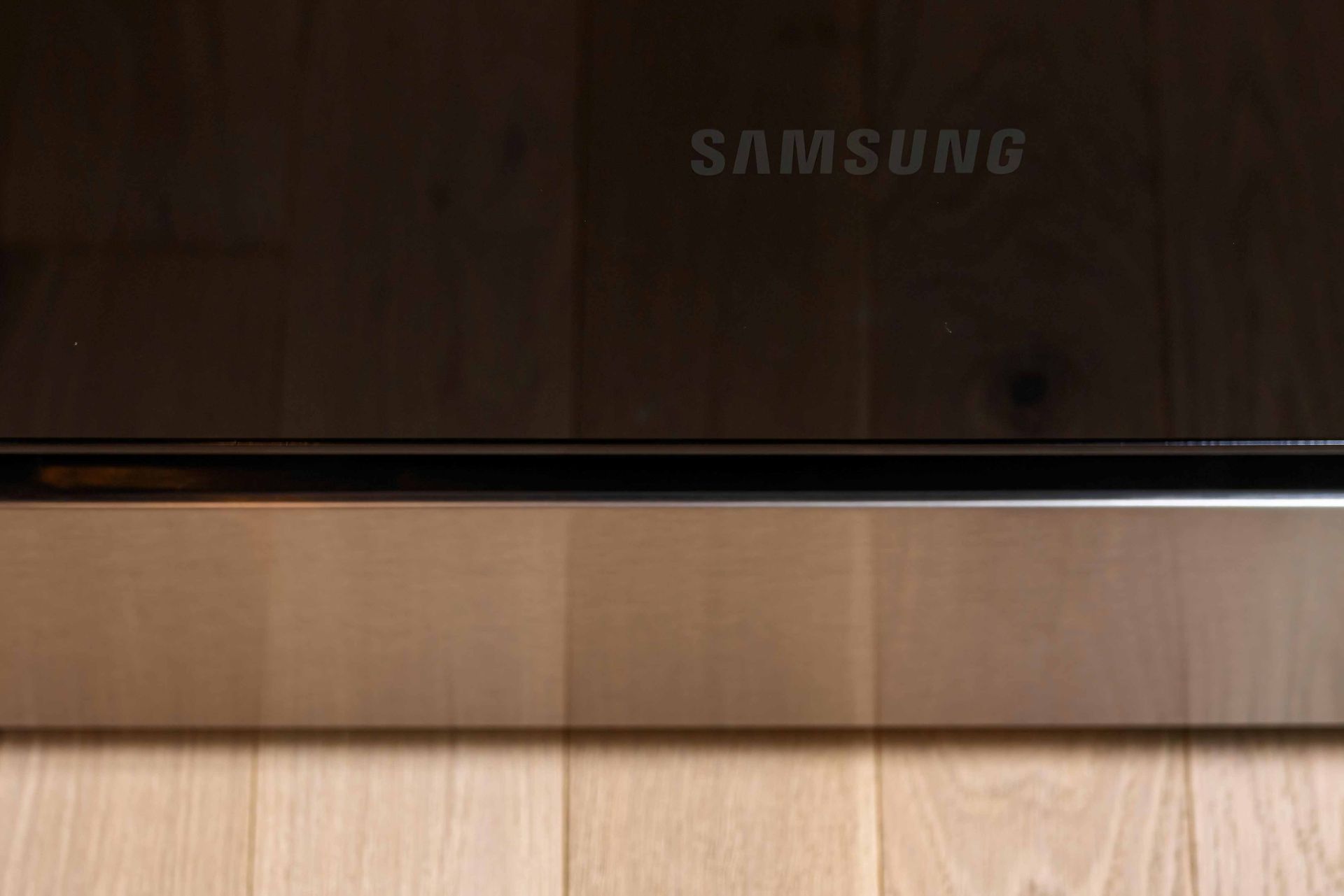 Black Samsung TV screen reflecting a room, with light wood flooring below