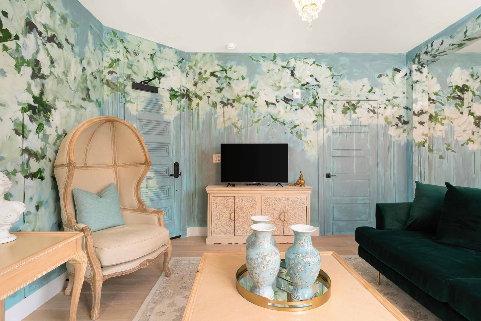 Pastel living room with floral mural, dark green sofa, rattan chair, TV, and decorative vases on a coffee table