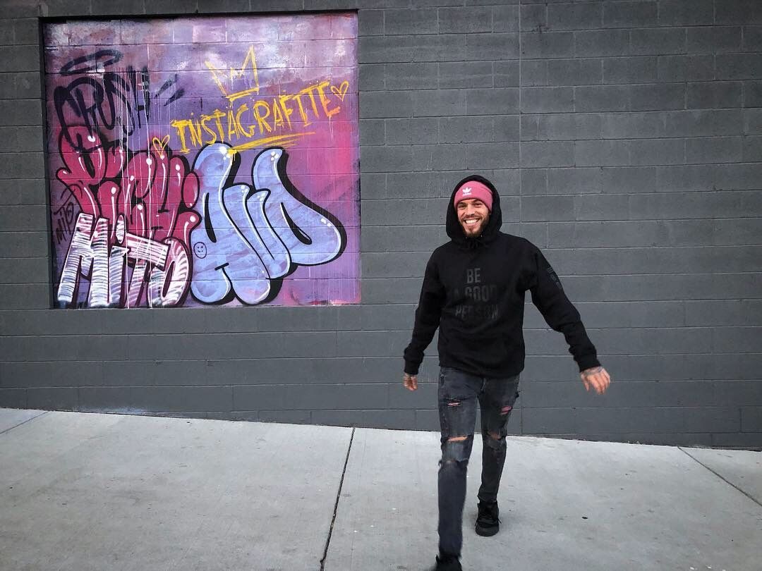Person in black hoodie and pink beanie walking beside a gray wall with colorful graffiti.