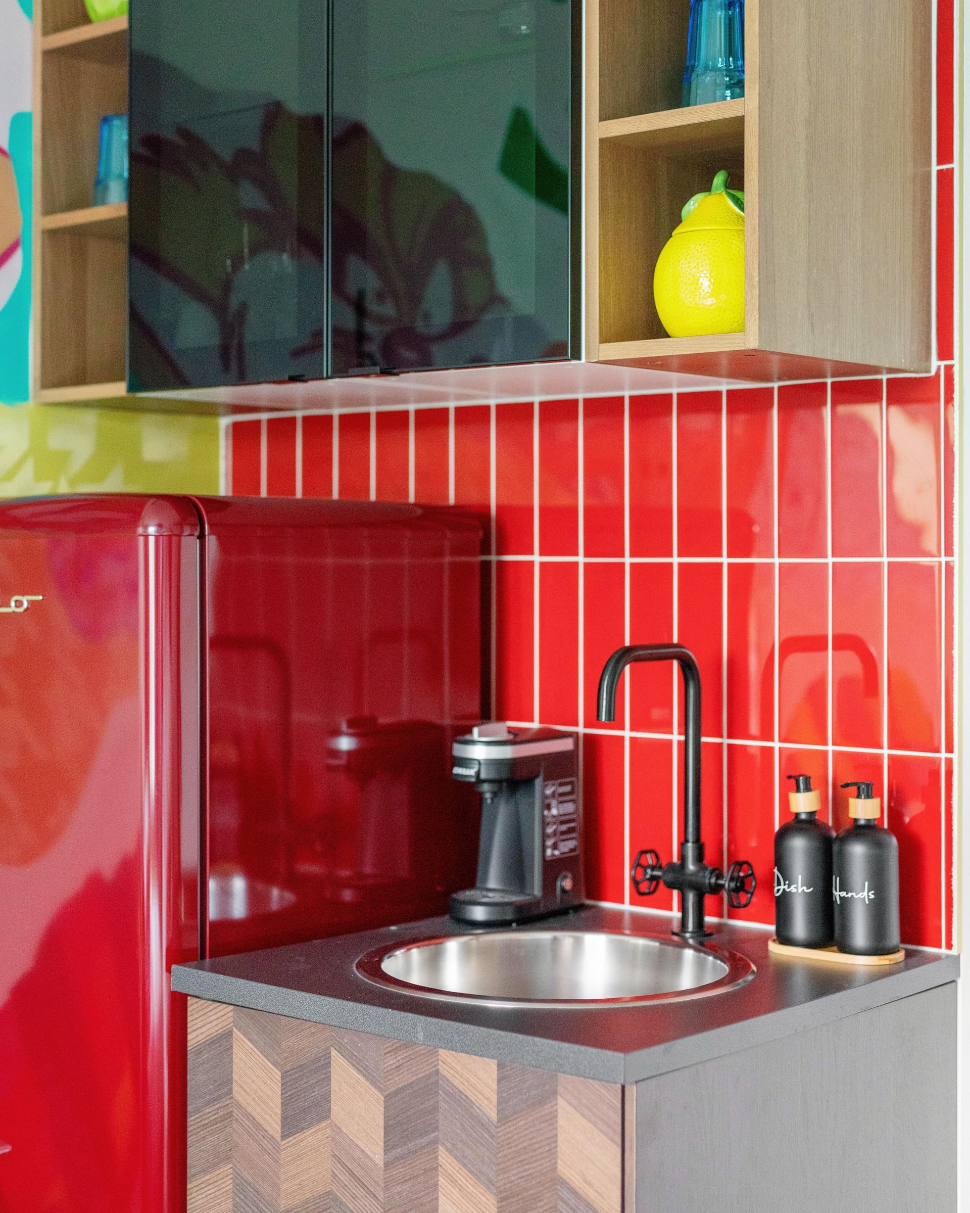 Compact red-tiled kitchen corner with sink, black faucet, red mini-fridge, and yellow kettle on shelf