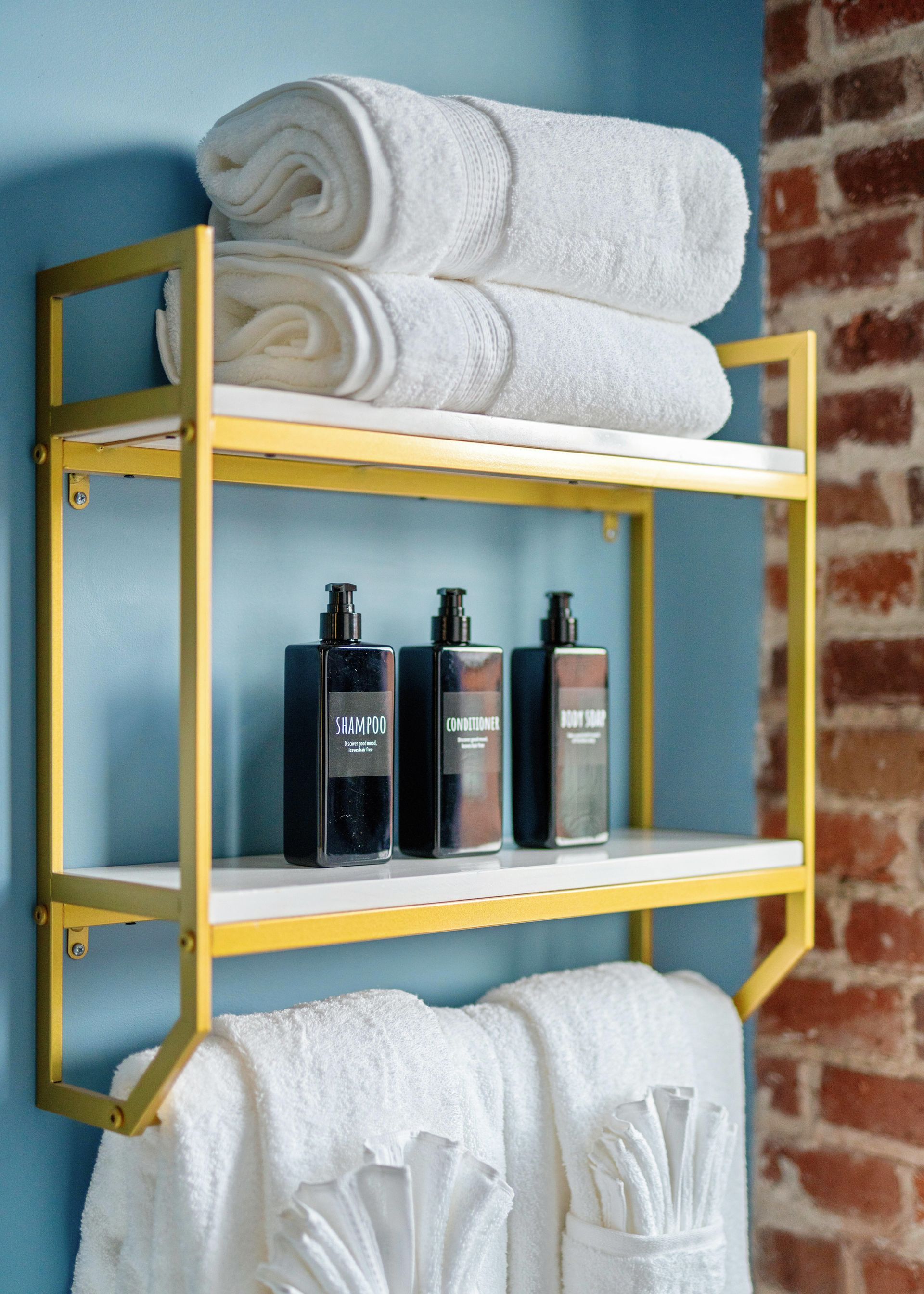 Yellow bathroom shelf with rolled white towels and dark bottles against a blue wall and brick accent