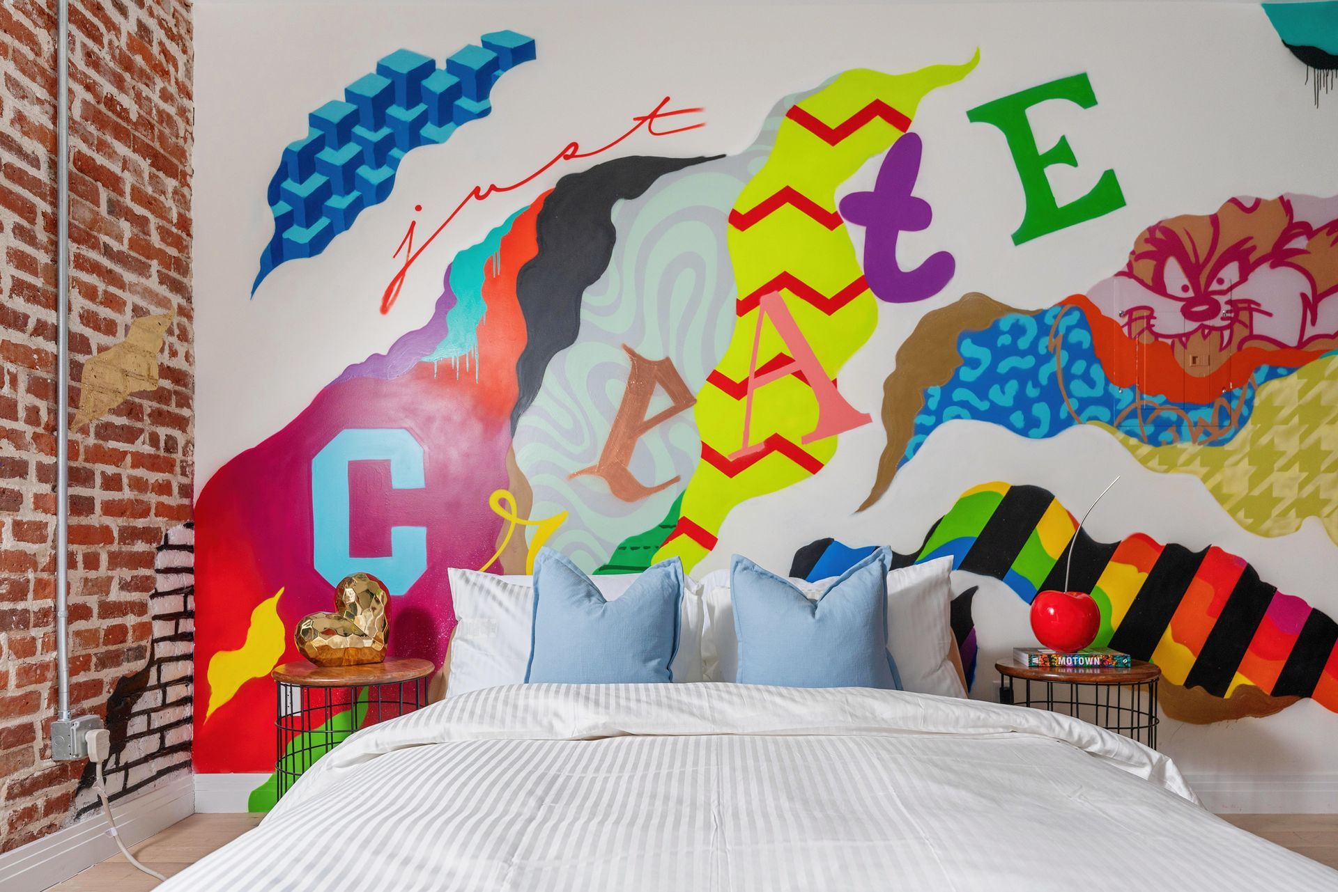 Colorful graffiti mural behind a bed in a modern bedroom with pillows and exposed brick walls