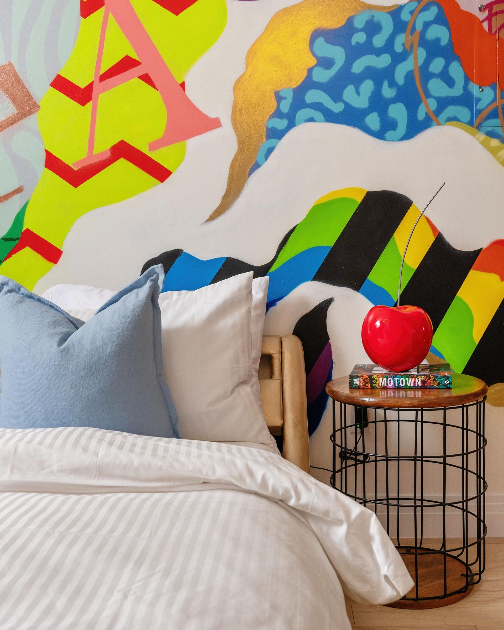 Colorful mural above a bed with blue pillows and a small wire side table holding a red ball lamp.