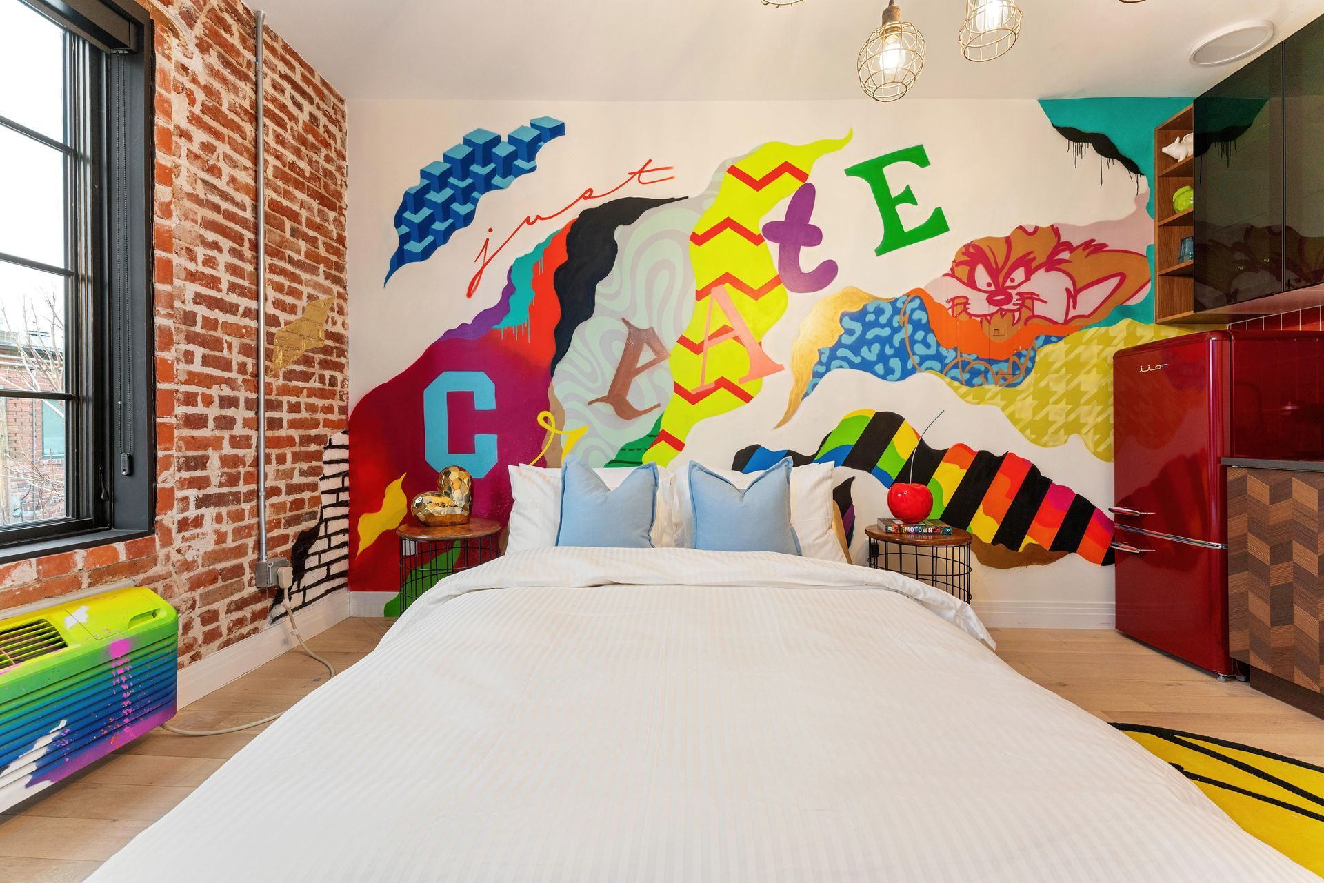 Colorful bedroom with graffiti mural, white bed, exposed brick wall, and red cabinets