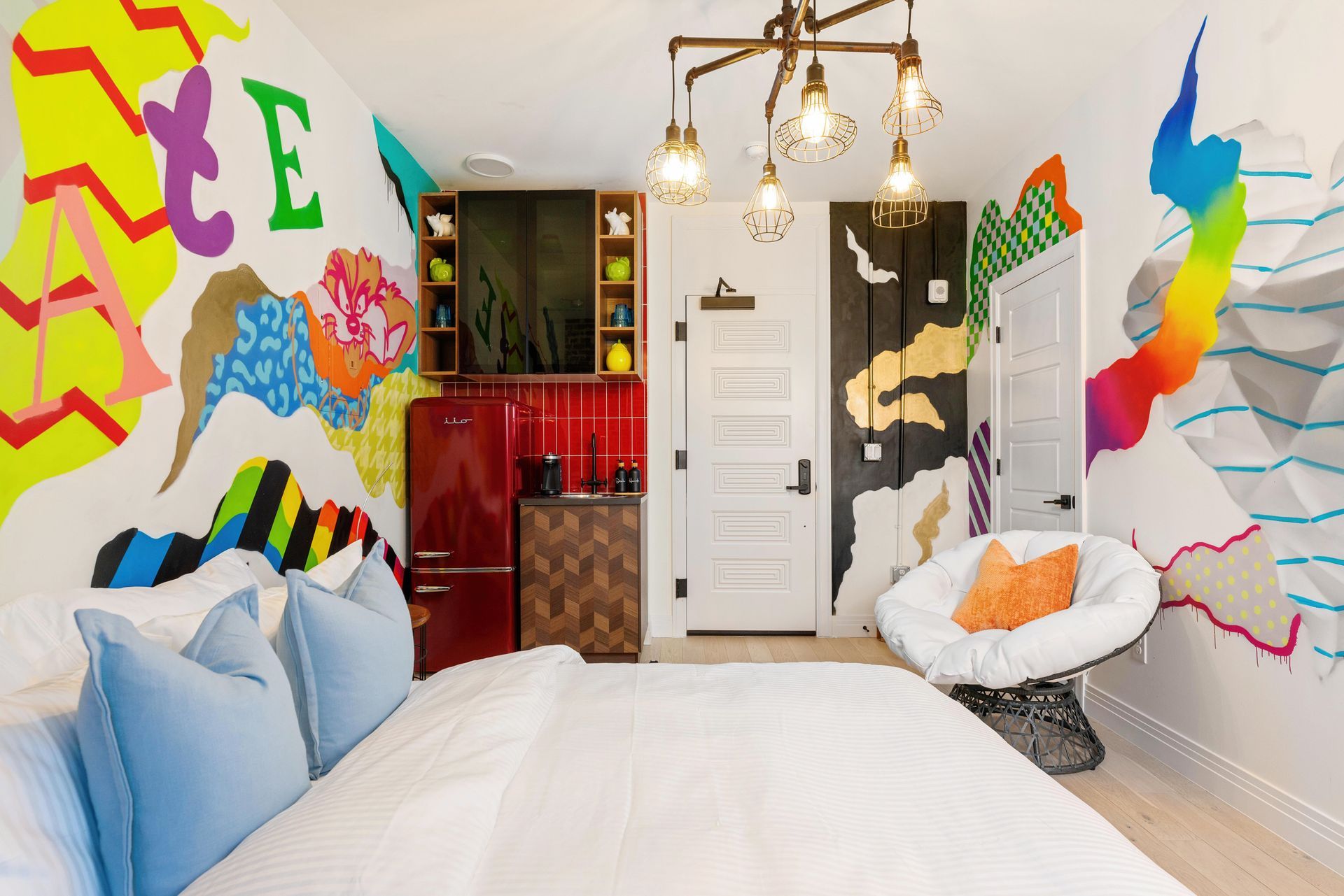 Colorful bedroom with abstract wall murals, a white bed, and a chair near a door and cabinet.