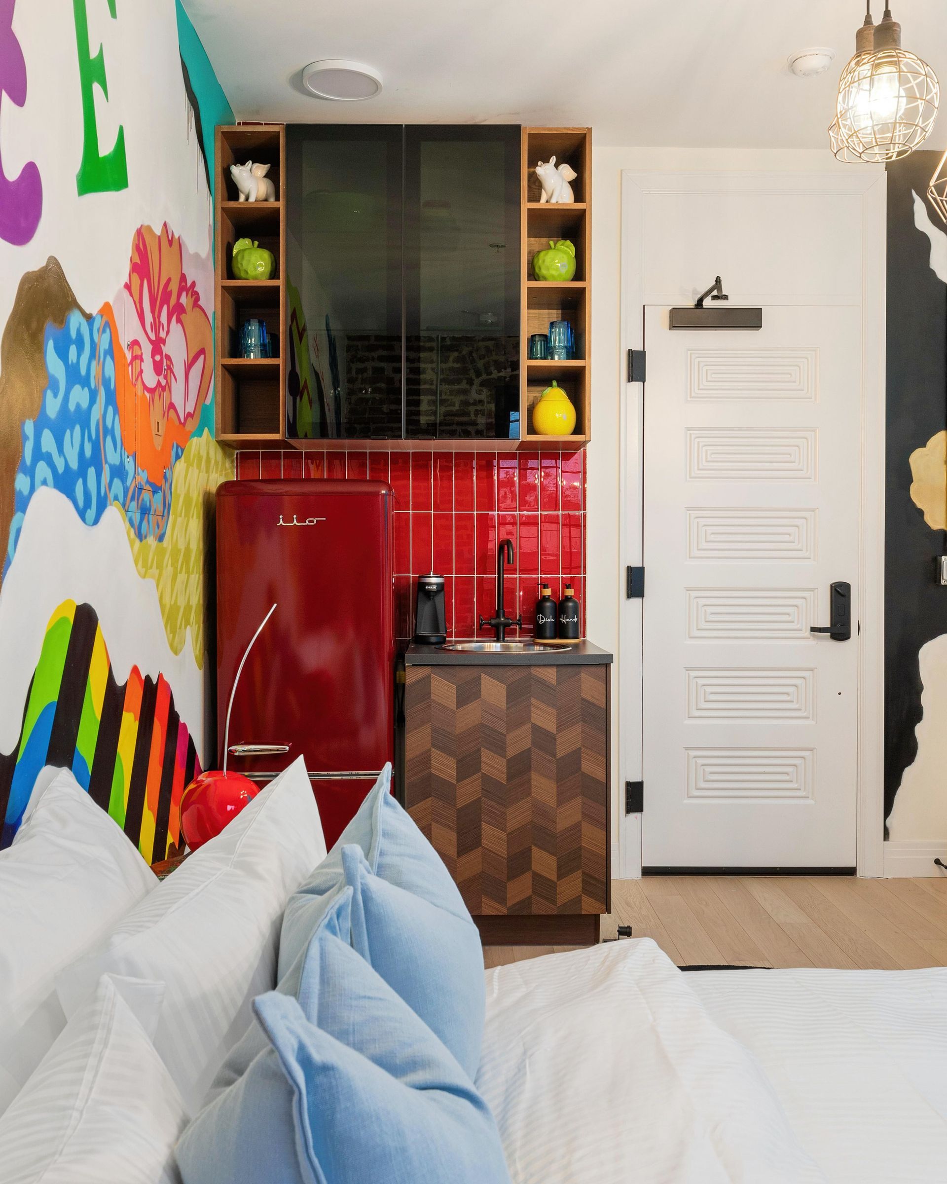 Colorful bedroom with white bed, blue pillows, red mini fridge, and compact kitchenette by a white door