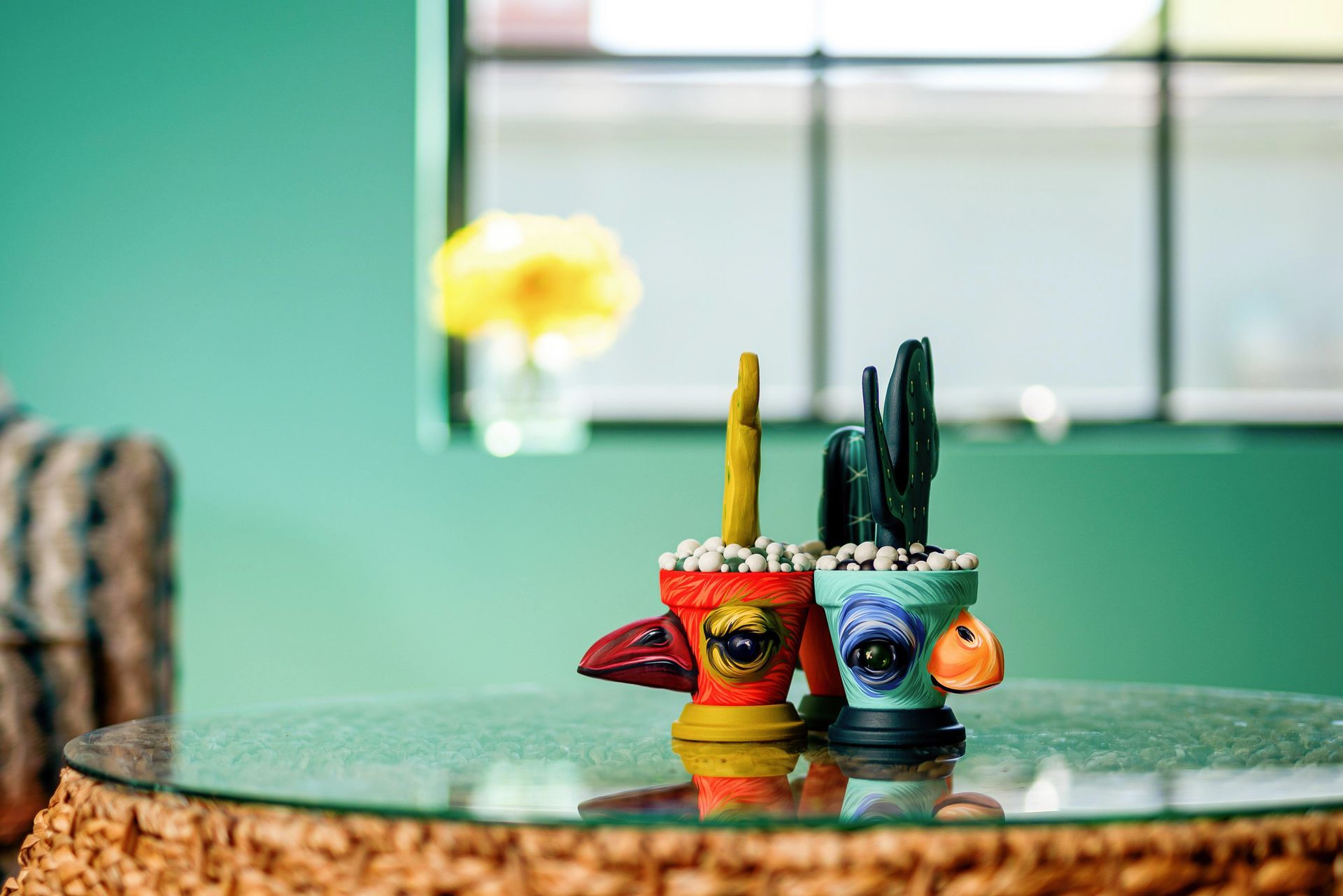 Two colorful mugs with cactus planters on a glass table in front of a teal wall and window