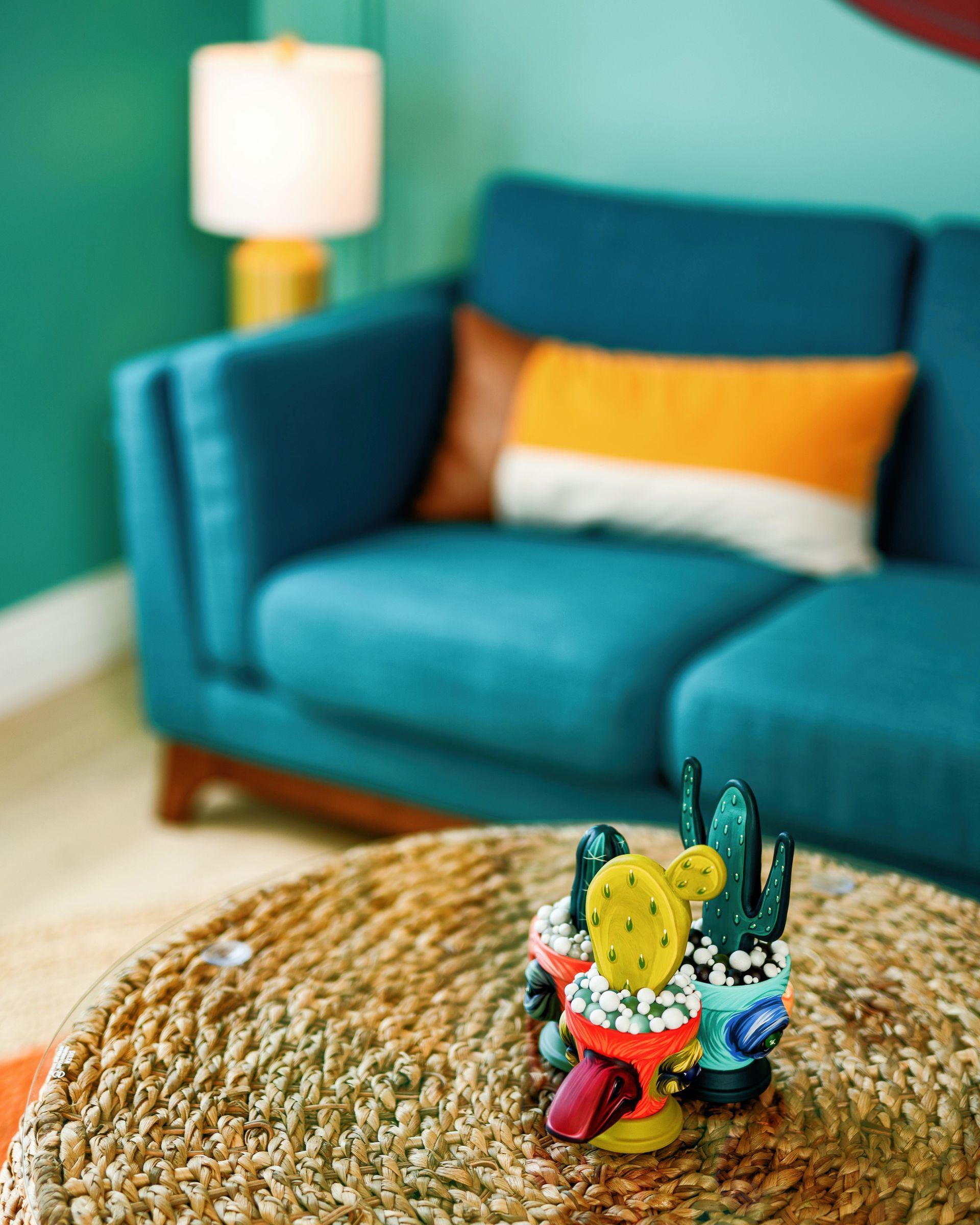 Small colorful cactus figurine on a woven table in a teal living room with a blue sofa and lamp.