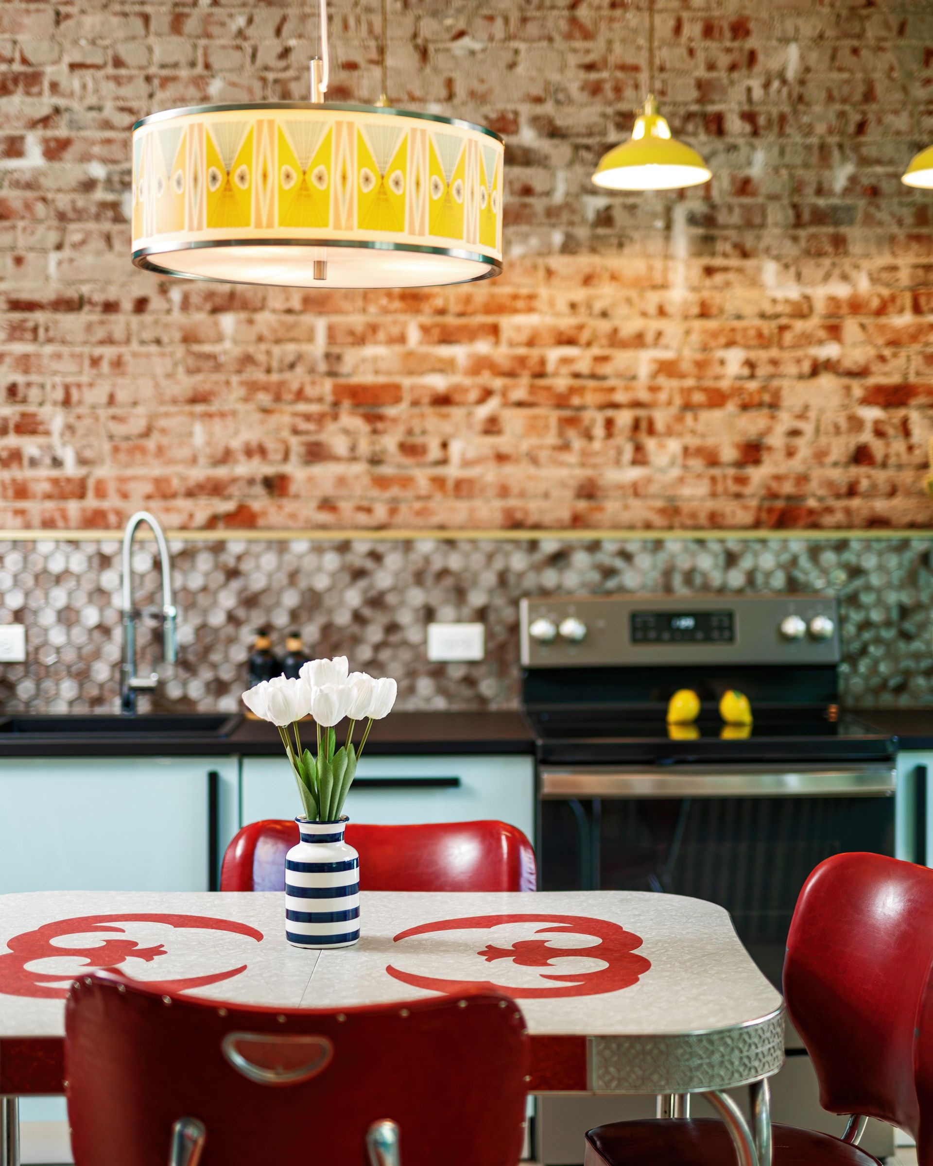 Modern kitchen with brick wall, yellow pendant lights, red chairs, and a table with flowers and a bowl of fruit
