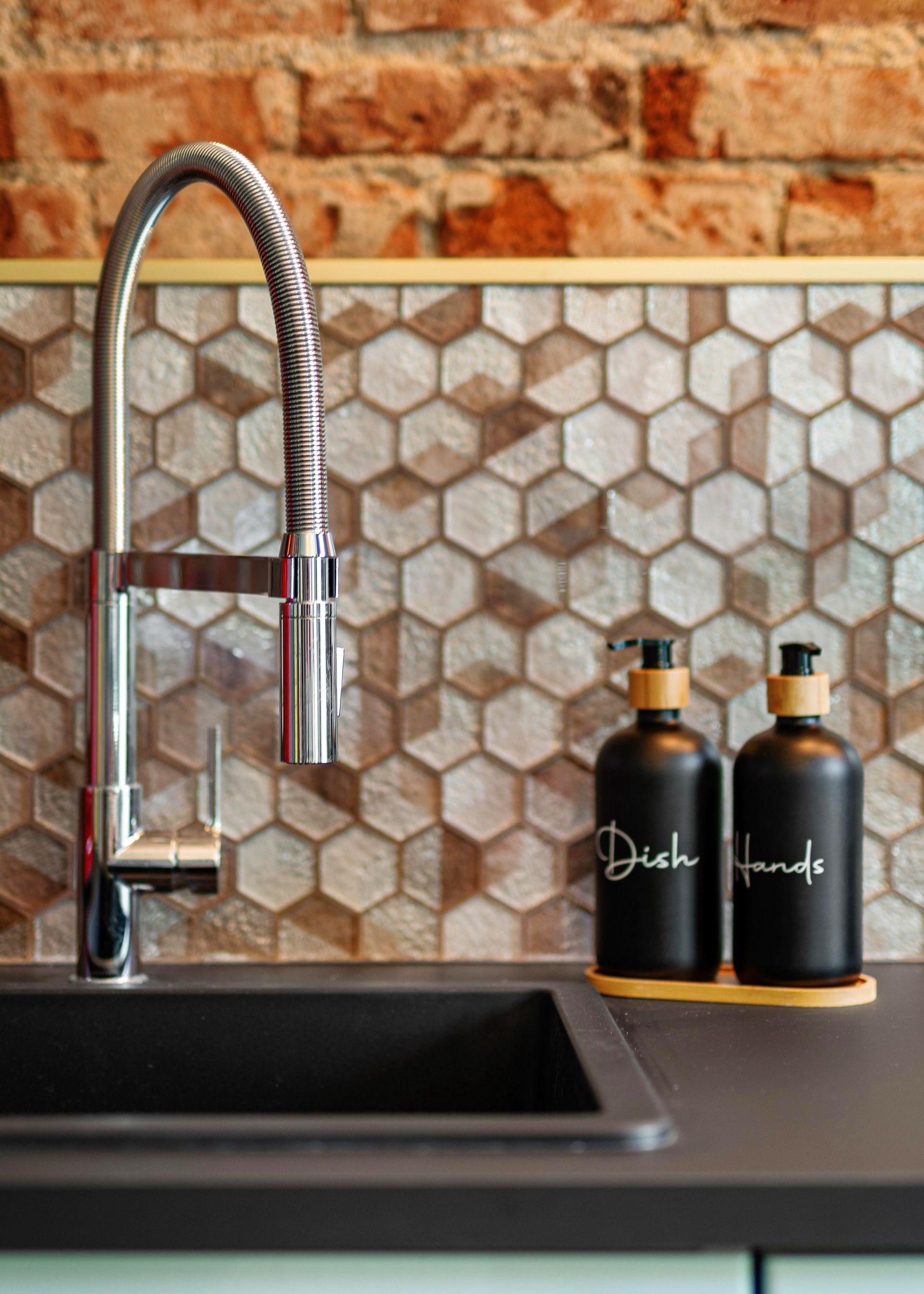 Black kitchen sink with chrome faucet beside two black soap bottles on a countertop against a tiled brick backsplash