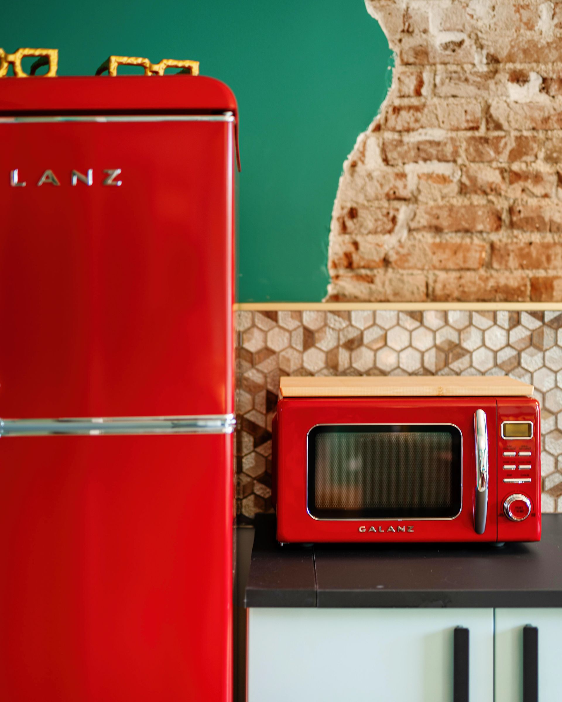 Red retro refrigerator and red microwave in a kitchen with green wall and exposed brick.
