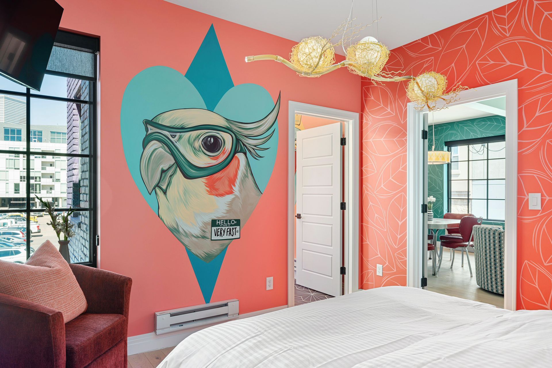 Colorful bedroom with coral walls, teal accent art, mirrored closet, and white bed in a bright city-view room