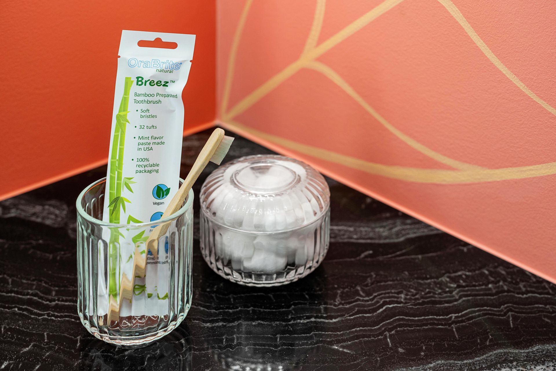 Toothbrushes in a glass beside cotton pads on a black shelf against a coral wall