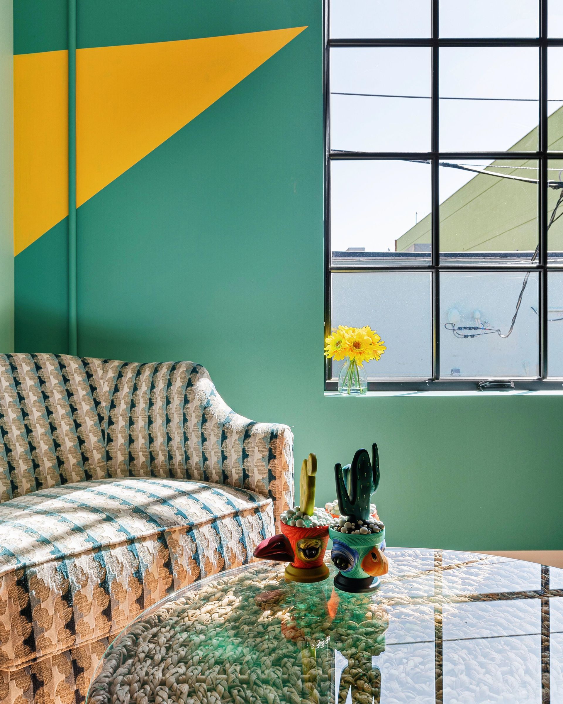 Cozy teal living room with patterned sofa, yellow wall accent, window, and glass coffee table with plants.