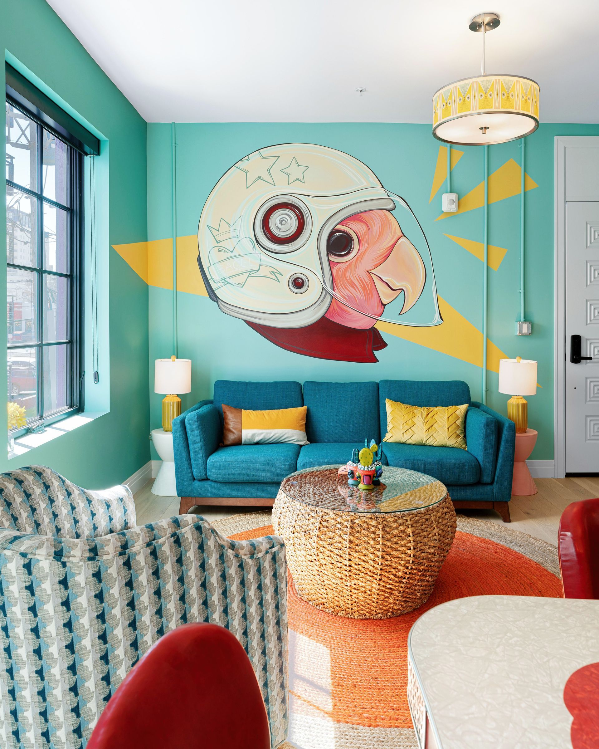 Bright teal living room with a large owl mural, teal sofa, yellow accents, and colorful chairs