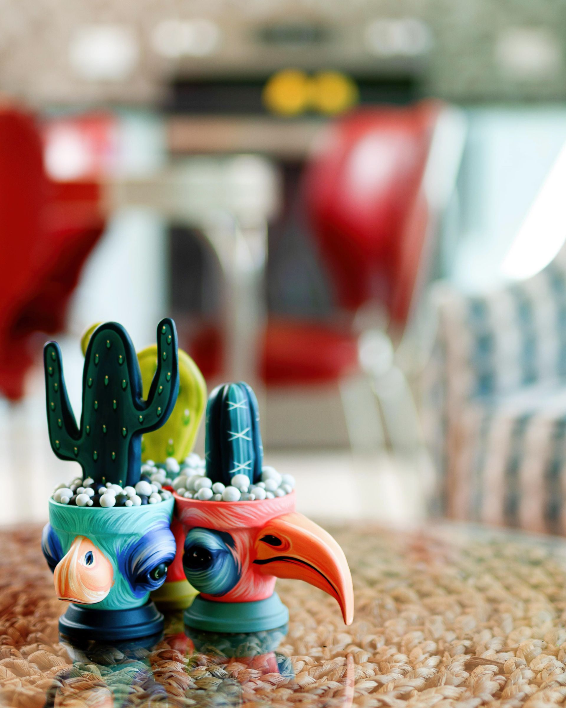 Two colorful ceramic bird figurines with cactus decorations on a woven table, blurred indoor background