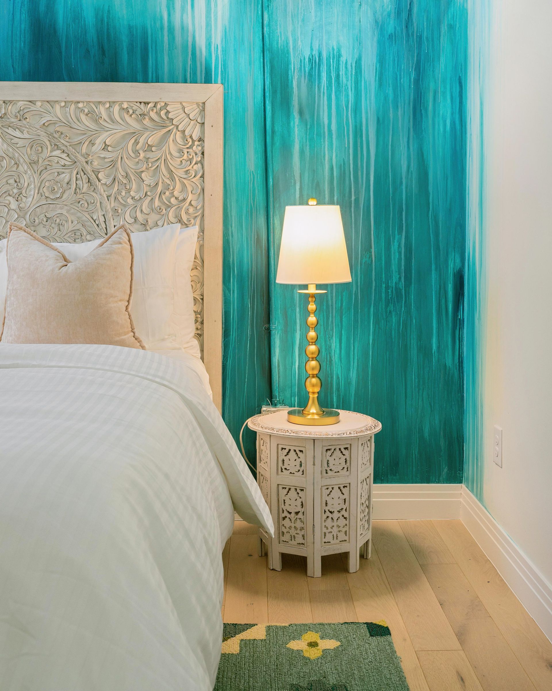 Turquoise bedroom corner with ornate bed, lamp on carved side table, and patterned rug.