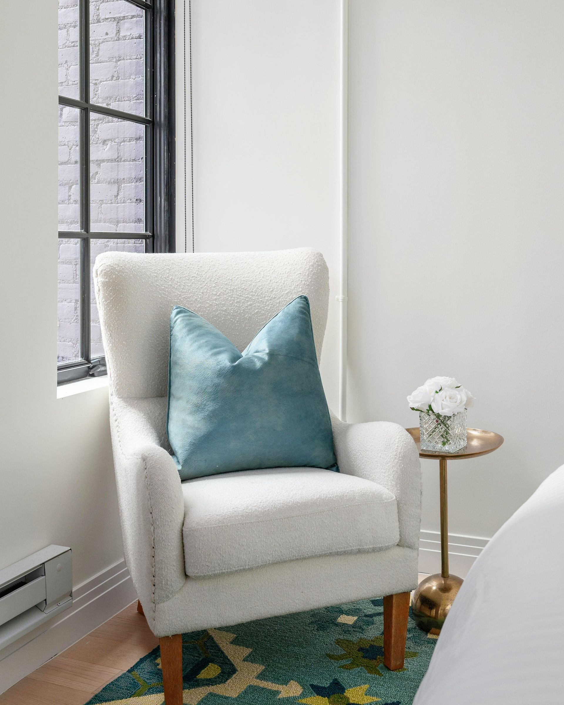 Cozy cream armchair with teal pillow beside a window, with a gold side table and floral arrangement.