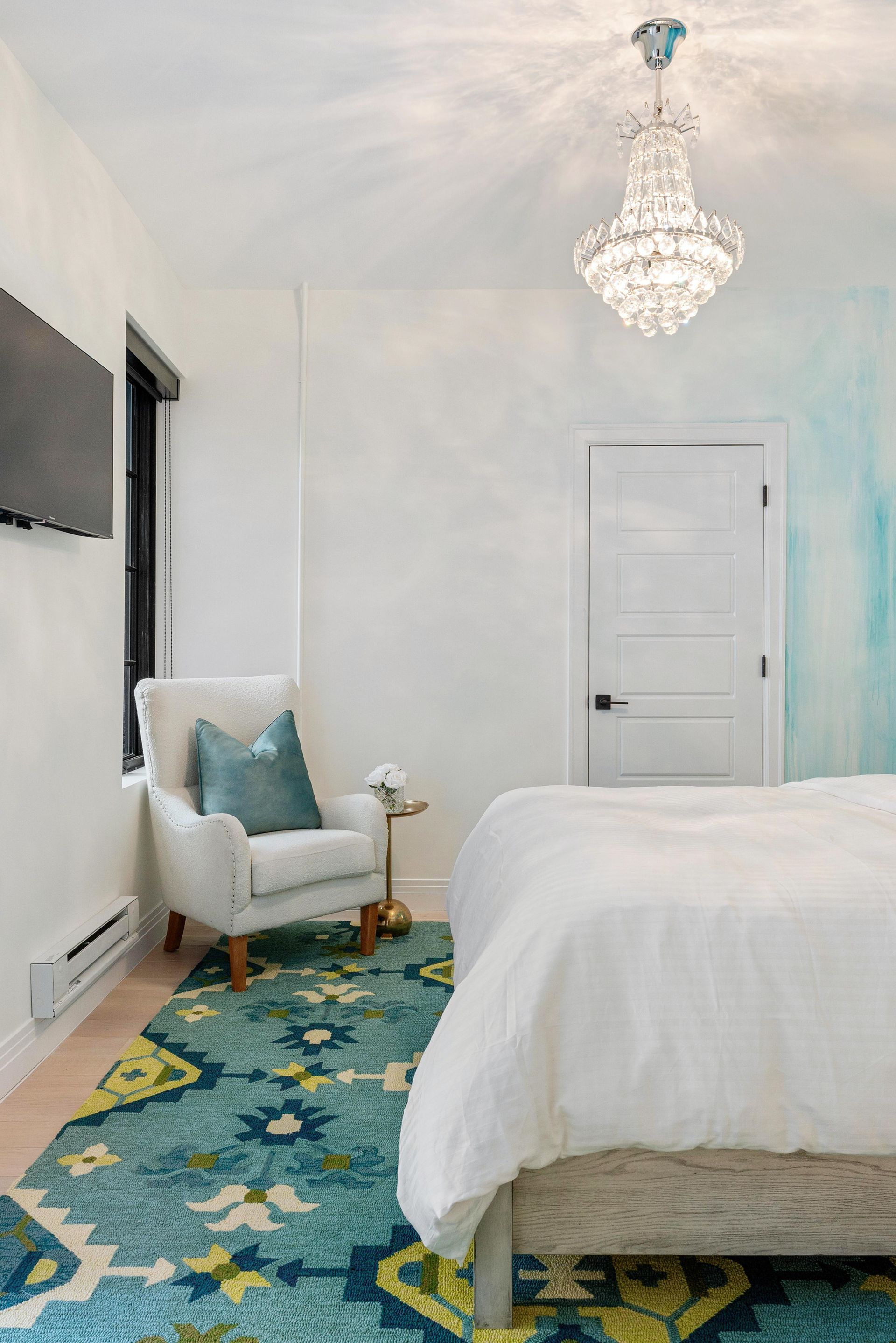 Elegant white bedroom with chandelier, teal patterned rug, armchair, and bed near a white door