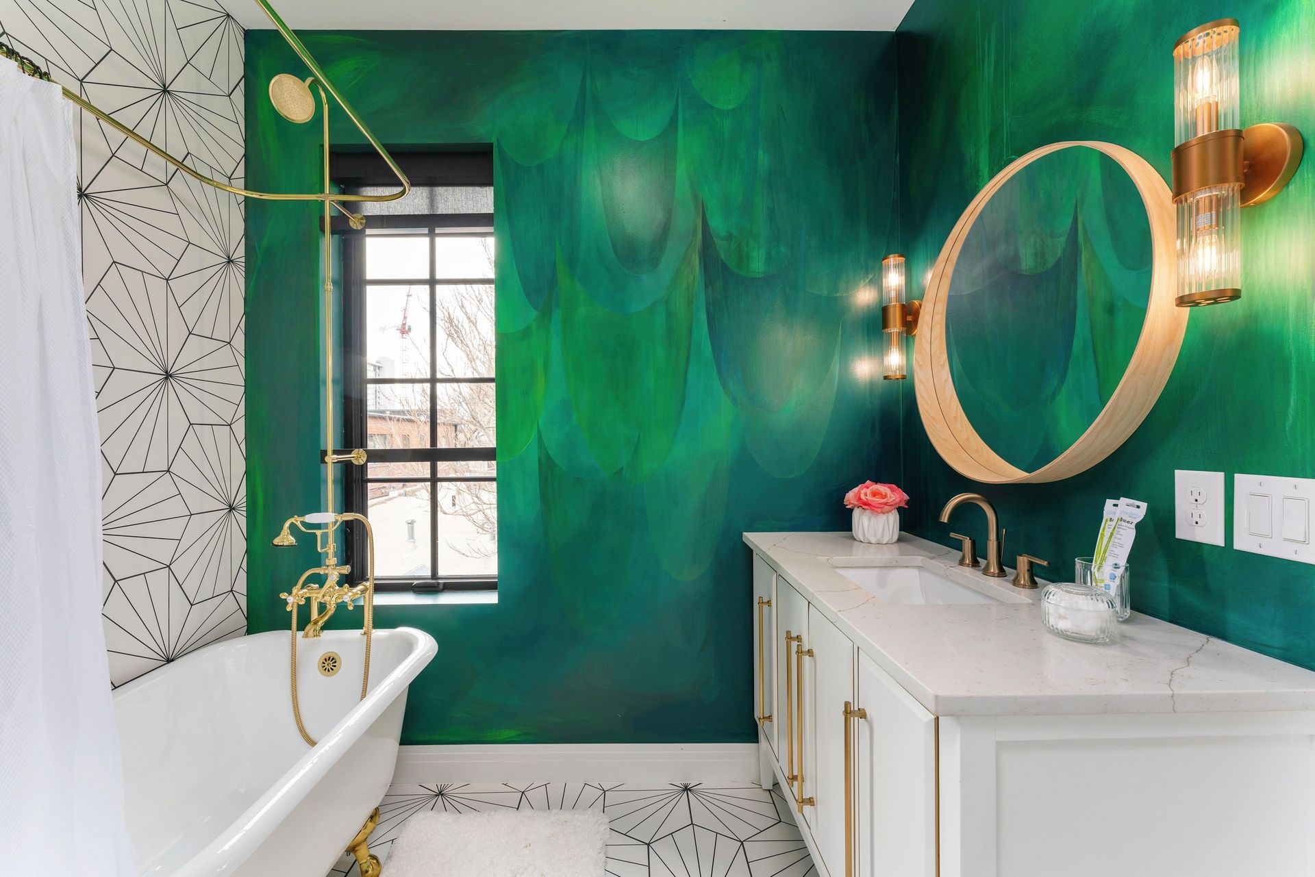 Bright bathroom with green mural wall, white vanity and tub, round mirror, and gold fixtures.