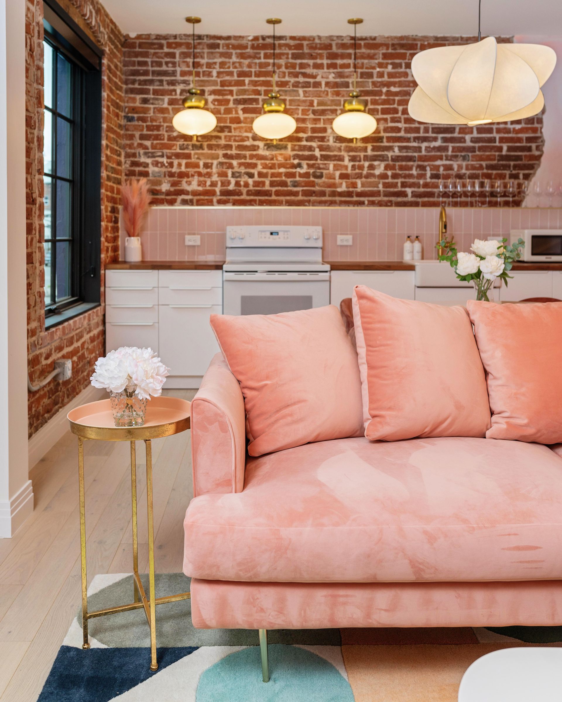 Pink sofa in a cozy kitchen with exposed brick, pendant lights, and a small gold side table.