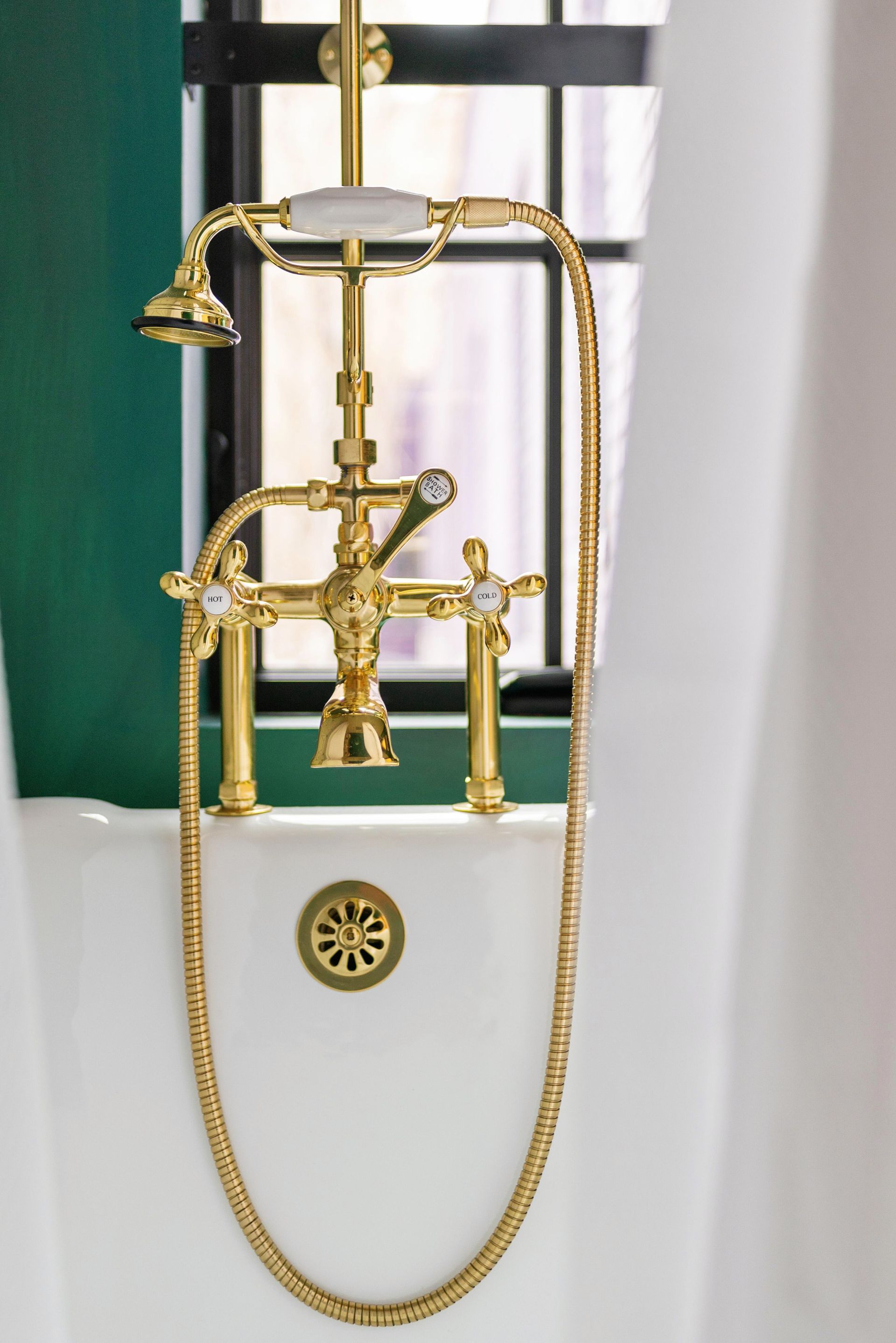 Gold freestanding bathtub faucet with handheld shower on a white tub against a green wall