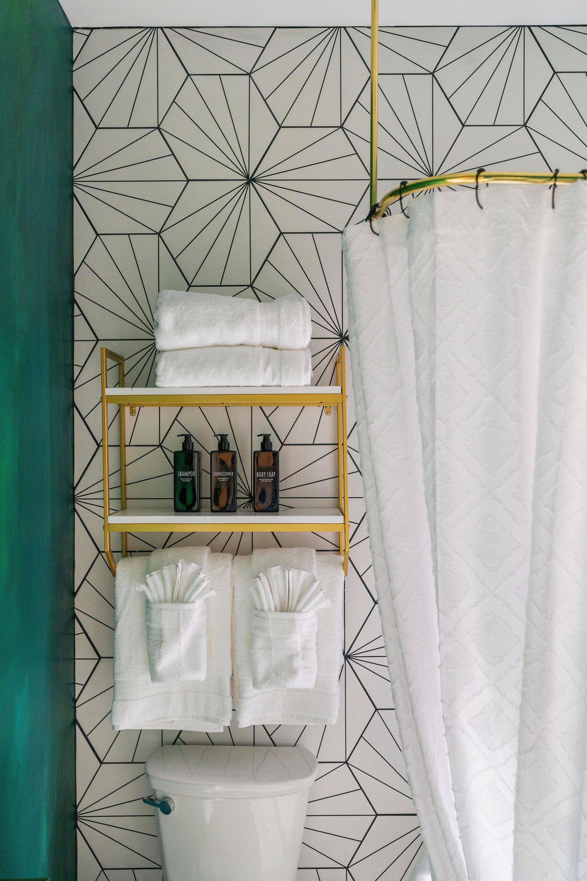 Small bathroom with black-and-white geometric wallpaper, yellow shelves, towels, toiletries, and a white shower curtain.
