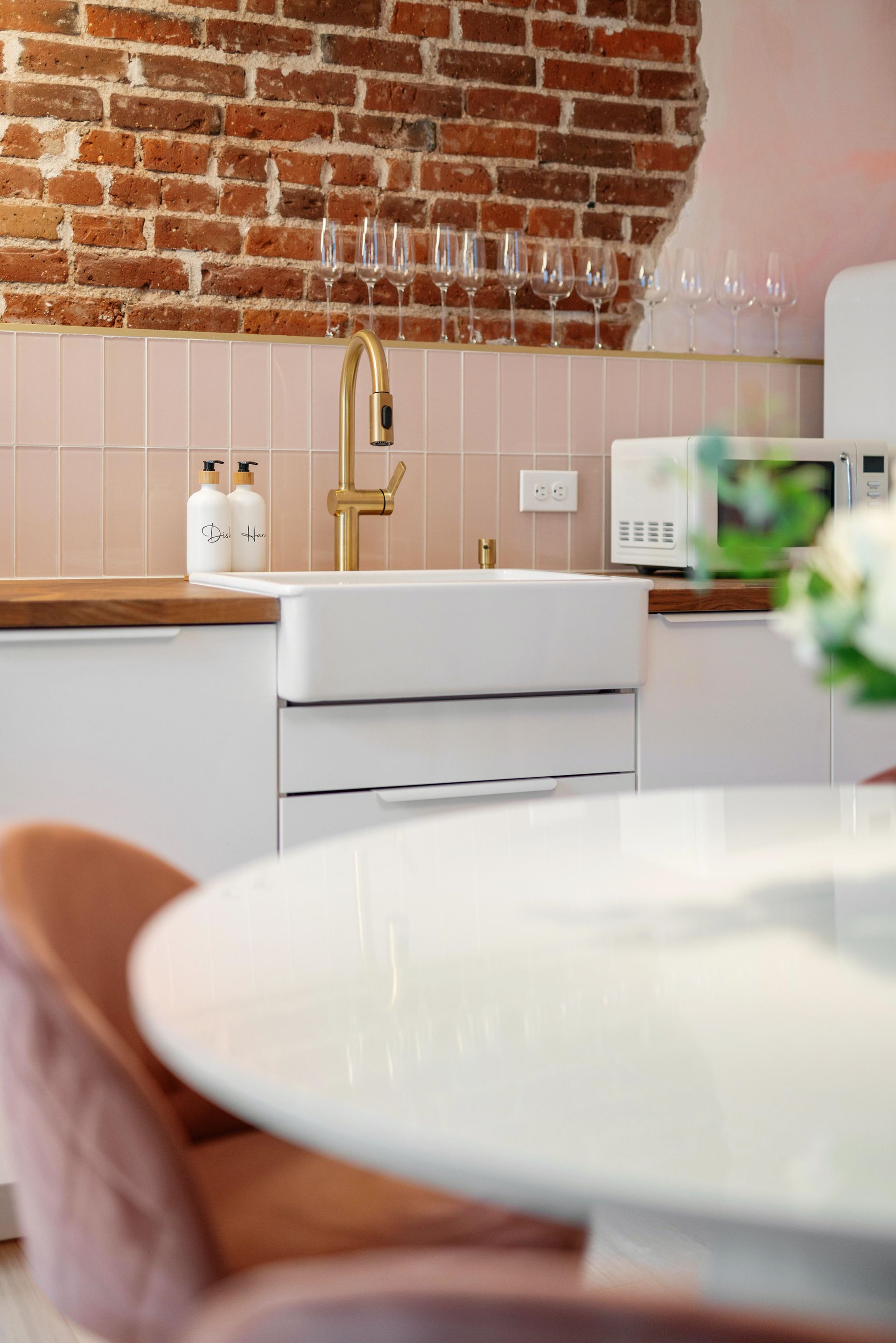 Modern kitchen with exposed brick wall, gold faucet, white sink, and round table in foreground