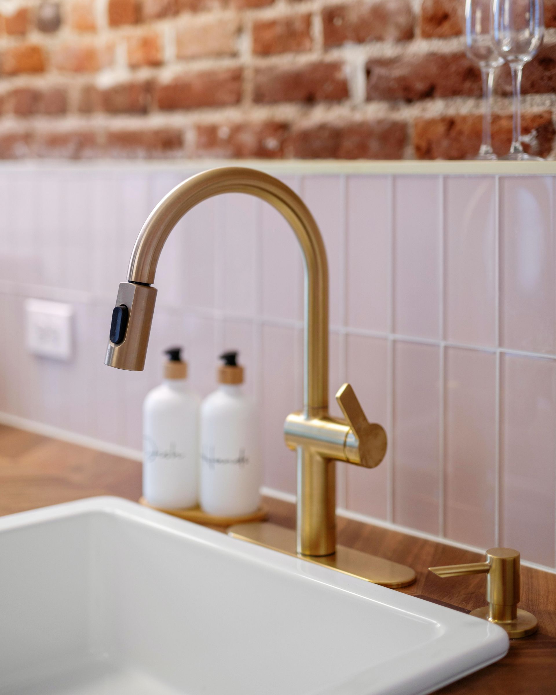 Brass kitchen faucet over white sink with pink tile backsplash and exposed brick wall