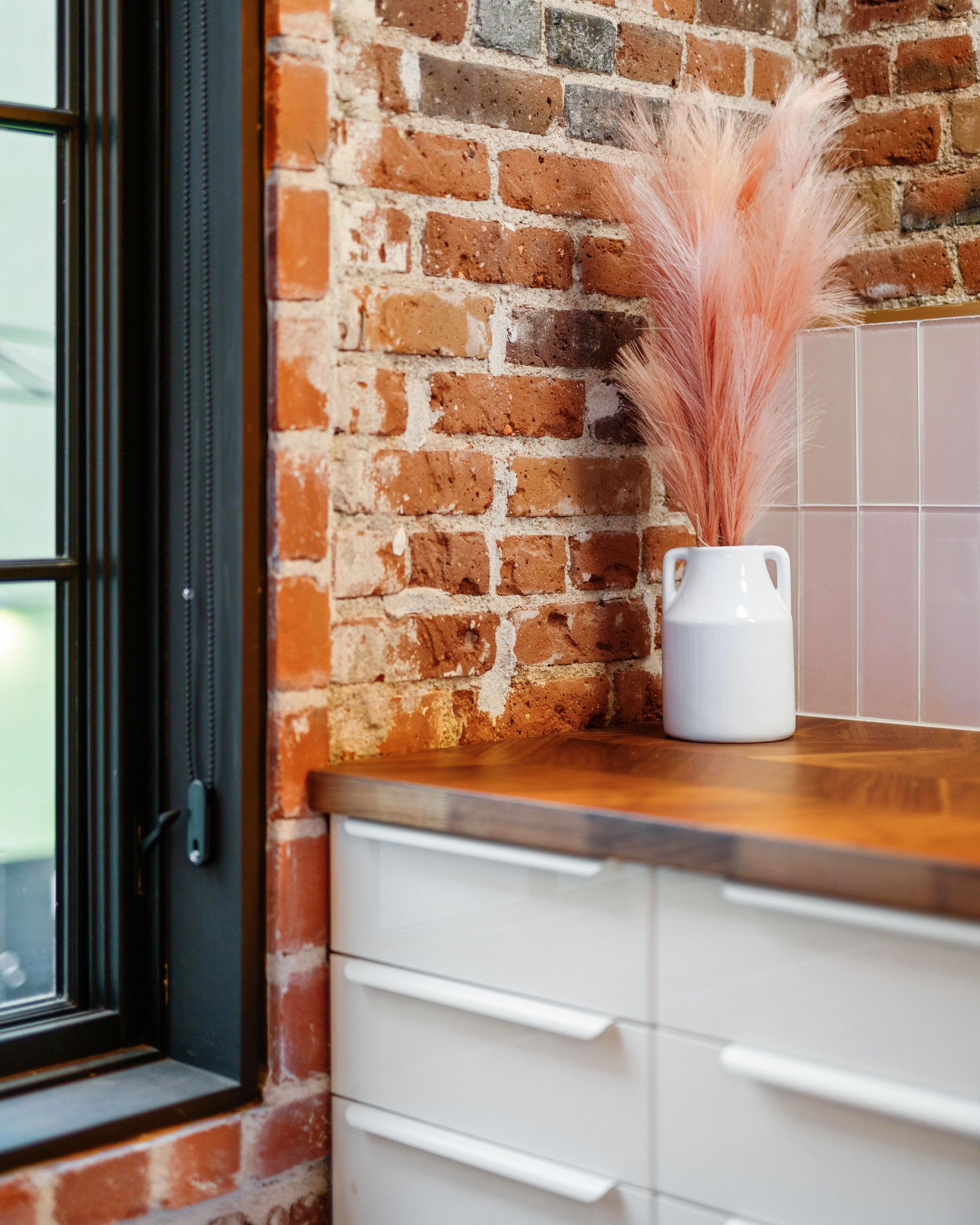 Brick kitchen corner with white cabinet, wood countertop, and vase of pink dried pampas grass by a window