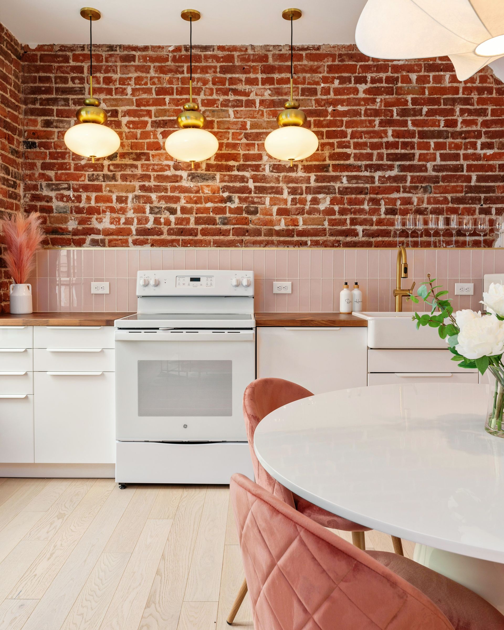Bright kitchen with white cabinets, exposed brick wall, white stove, and round table with pink chairs