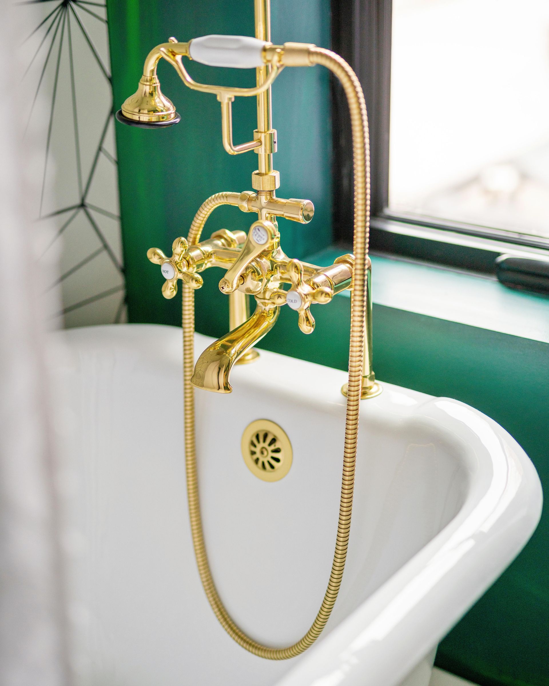 Gold vintage bathtub faucet over a white tub with green tile wall and window light