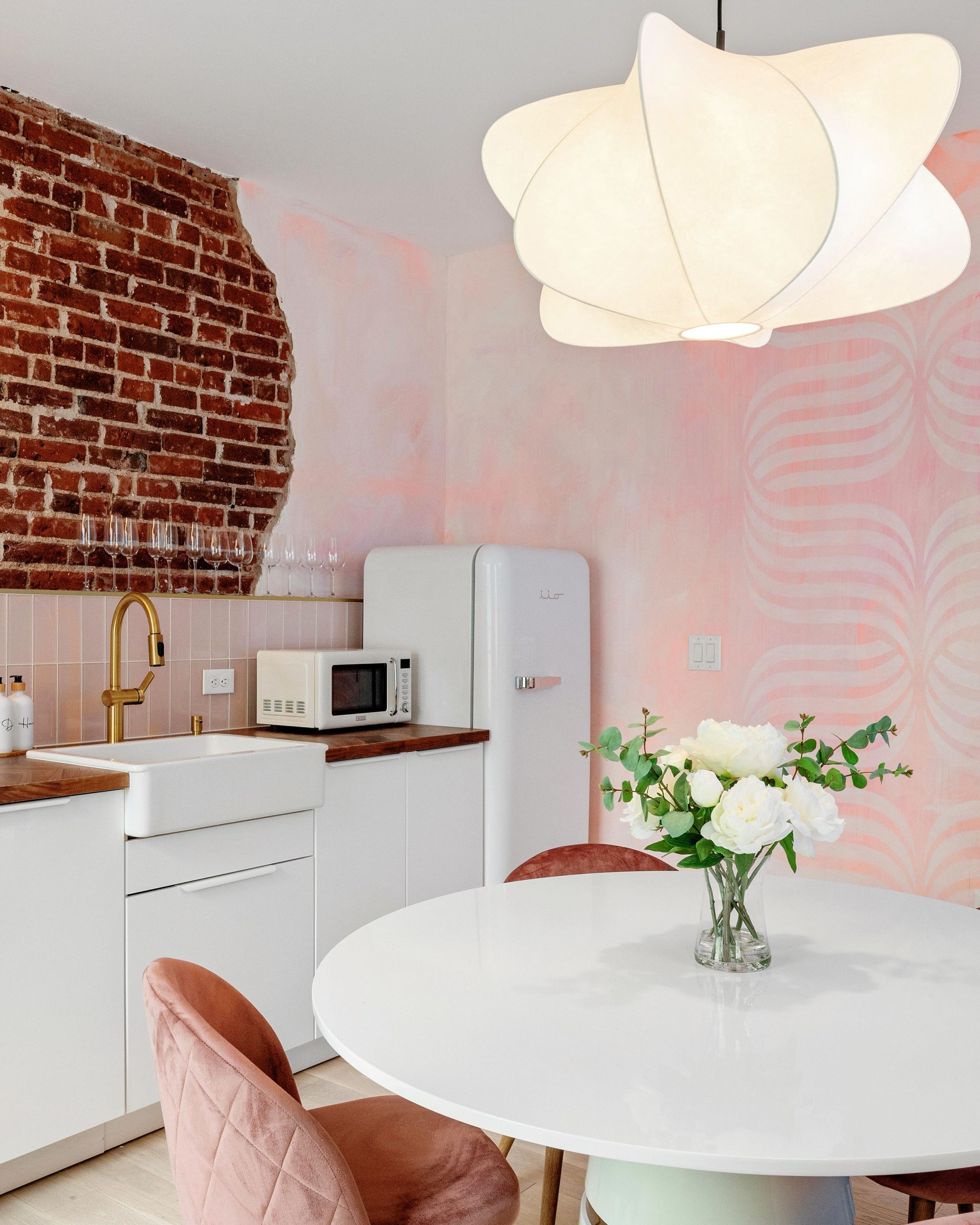 Bright kitchen with white table, flowers, pink walls, exposed brick, and hanging white pendant light