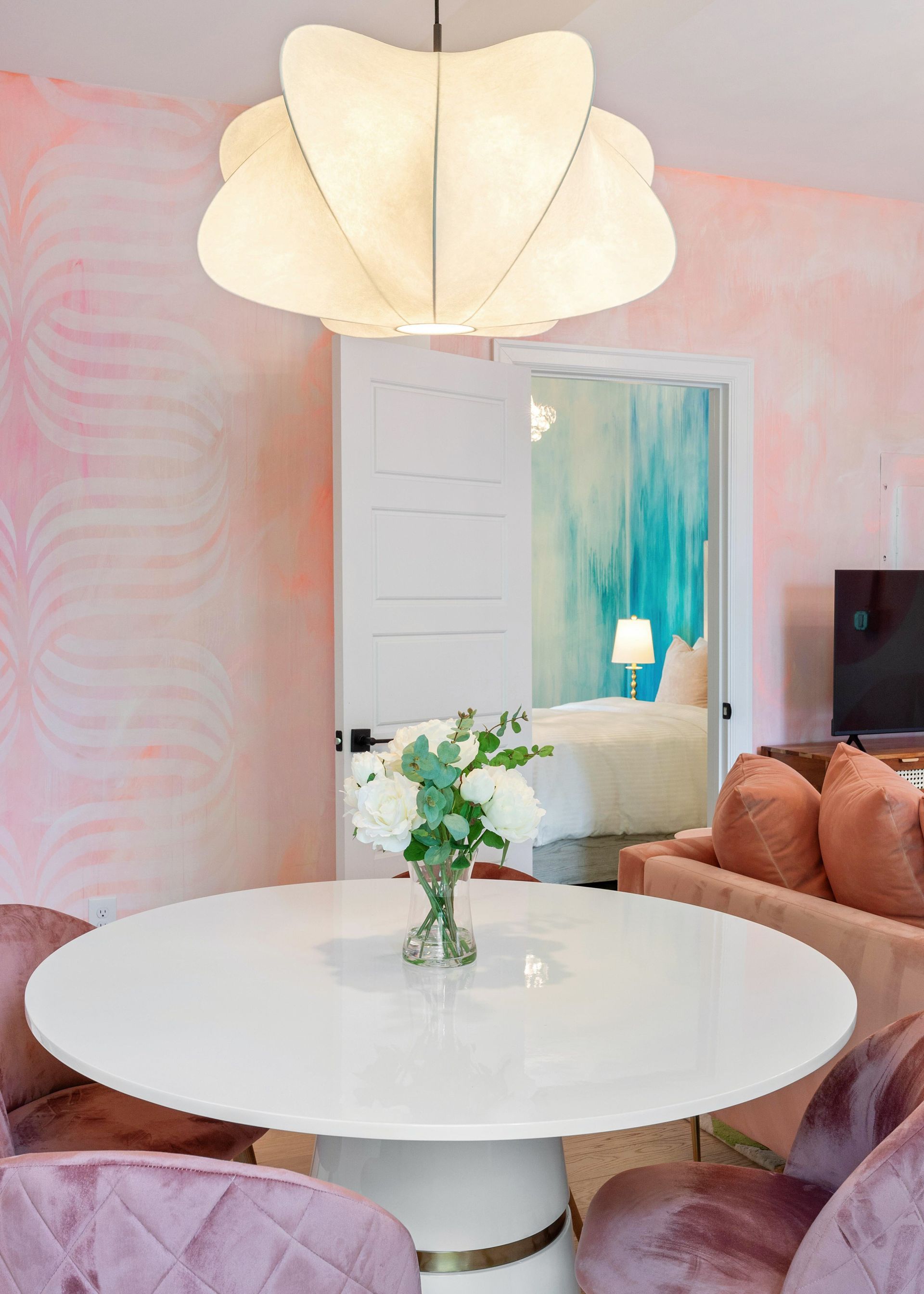 Bright dining nook with a round white table, floral centerpiece, and pink walls under a large pendant light