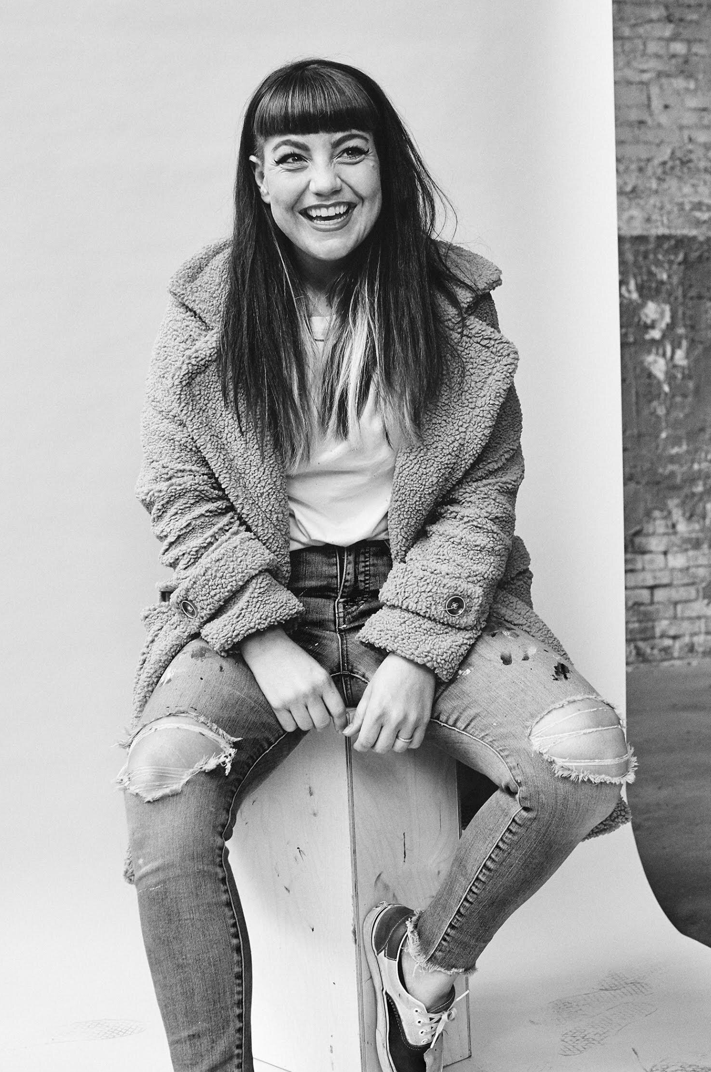 Person sitting on a box, smiling, in a fur coat and ripped jeans against a plain wall.