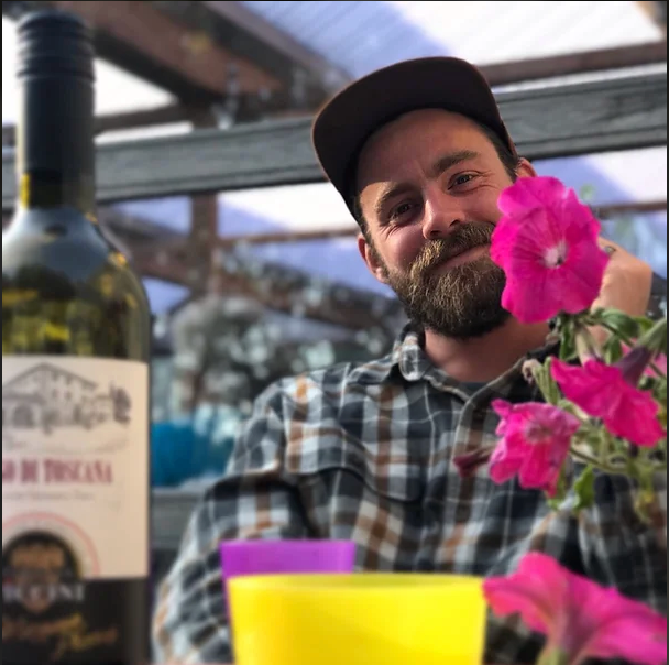 Smiling bearded man in a cap sits by pink flowers with a wine bottle and colorful cups on the table