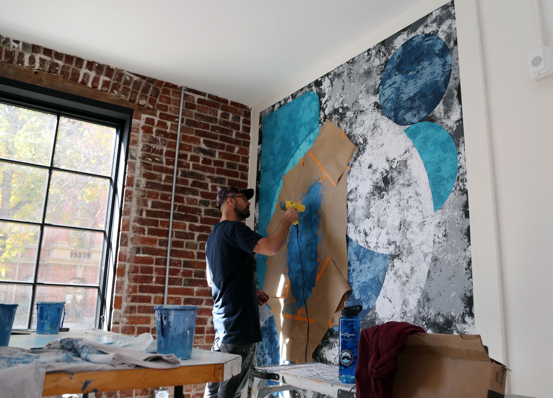 Person painting a large abstract mural on a brick wall in a bright room with windows.