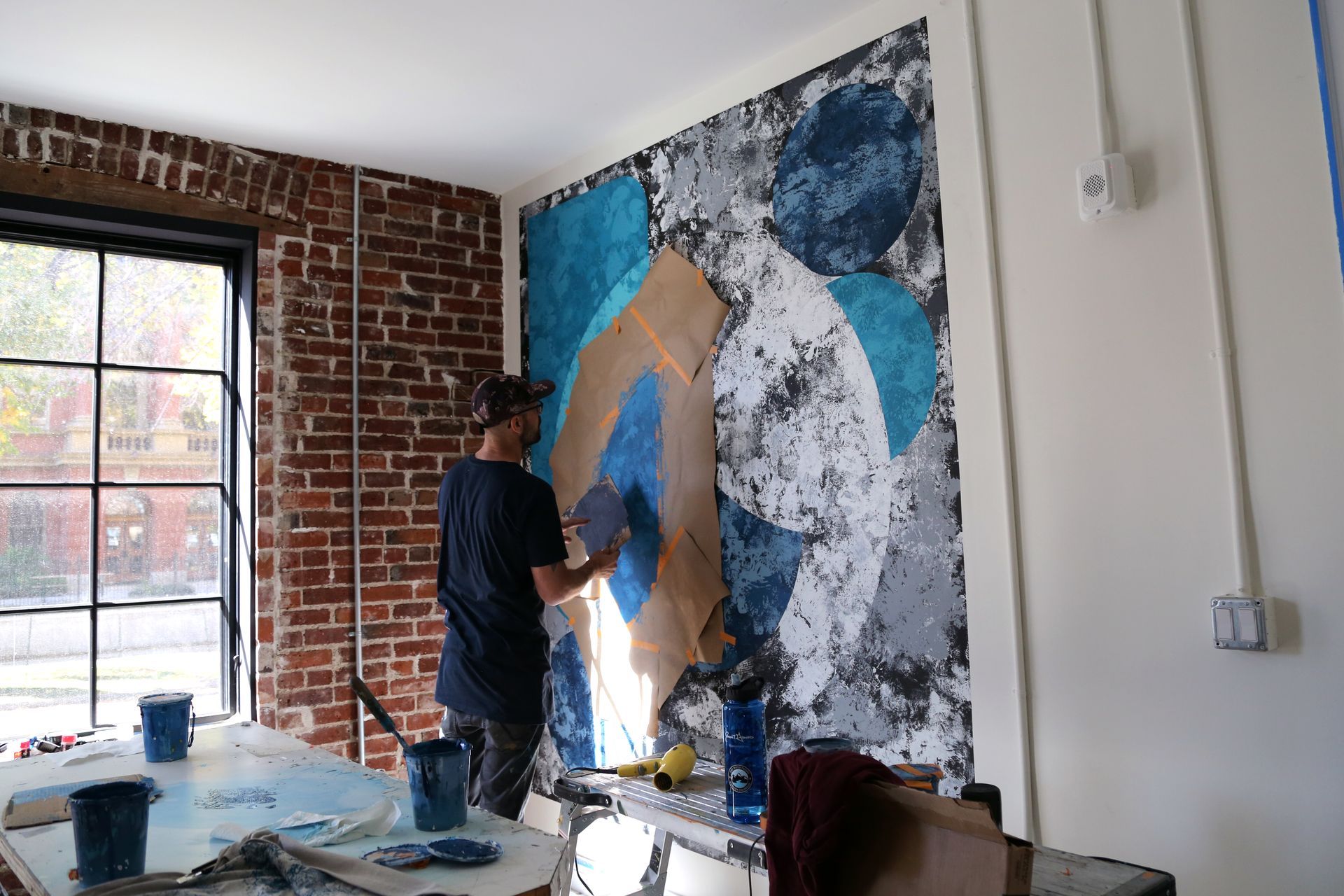 Artist painting a large abstract mural on an interior brick wall beside a window.
