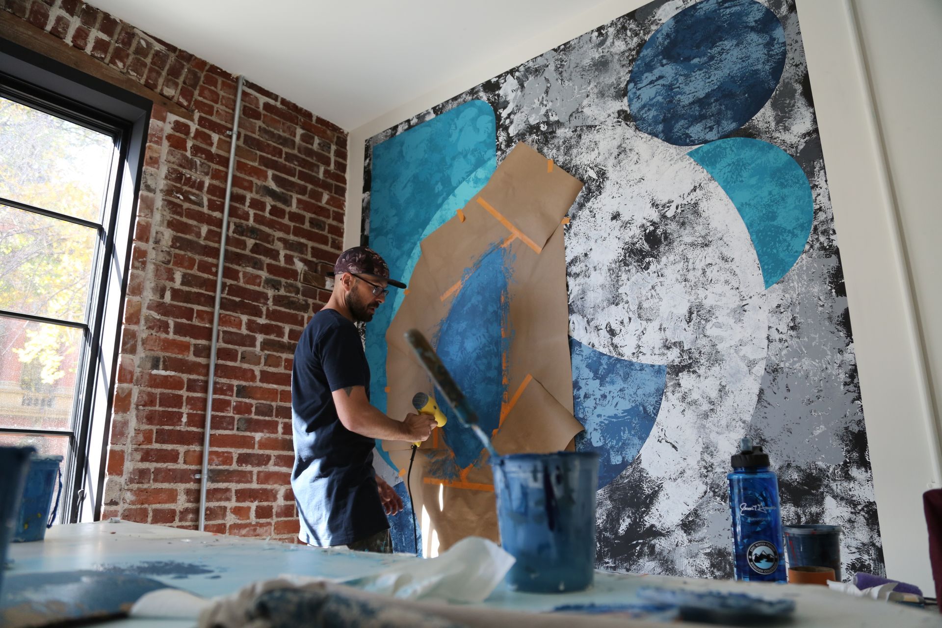 Person painting a large abstract mural on a brick wall in a bright room
