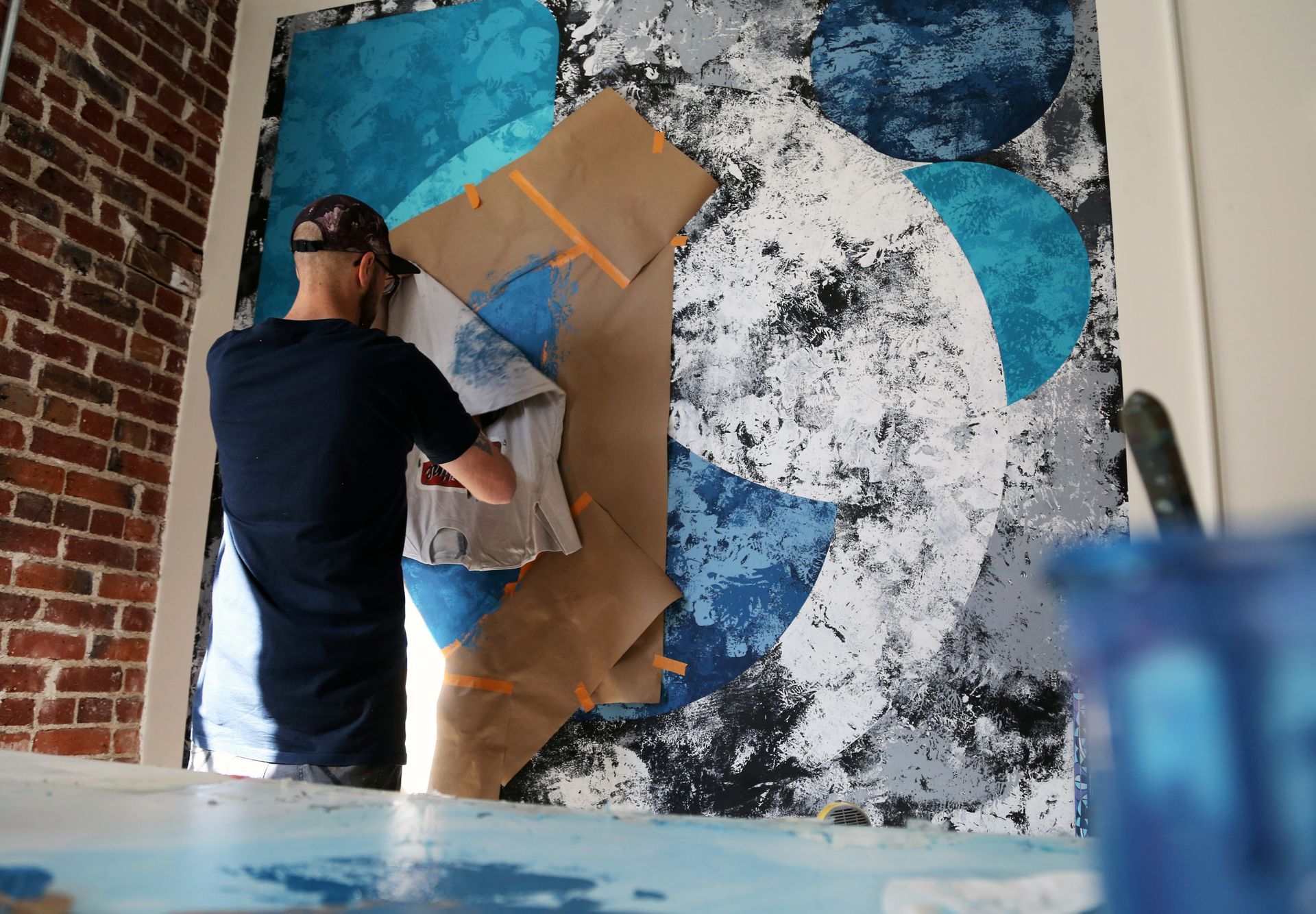 Person painting a large abstract mural with blue, white, black, and tan shapes on a brick wall