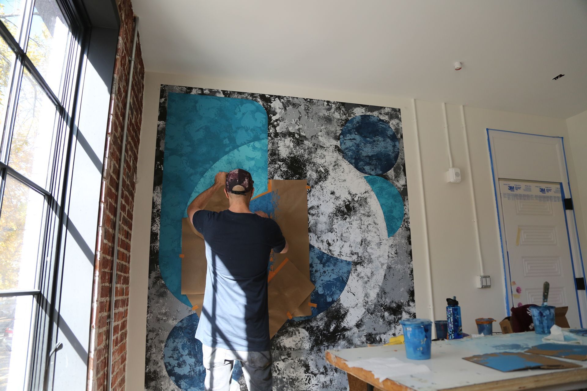 Artist painting a large abstract mural on a studio wall beside a sunlit window.