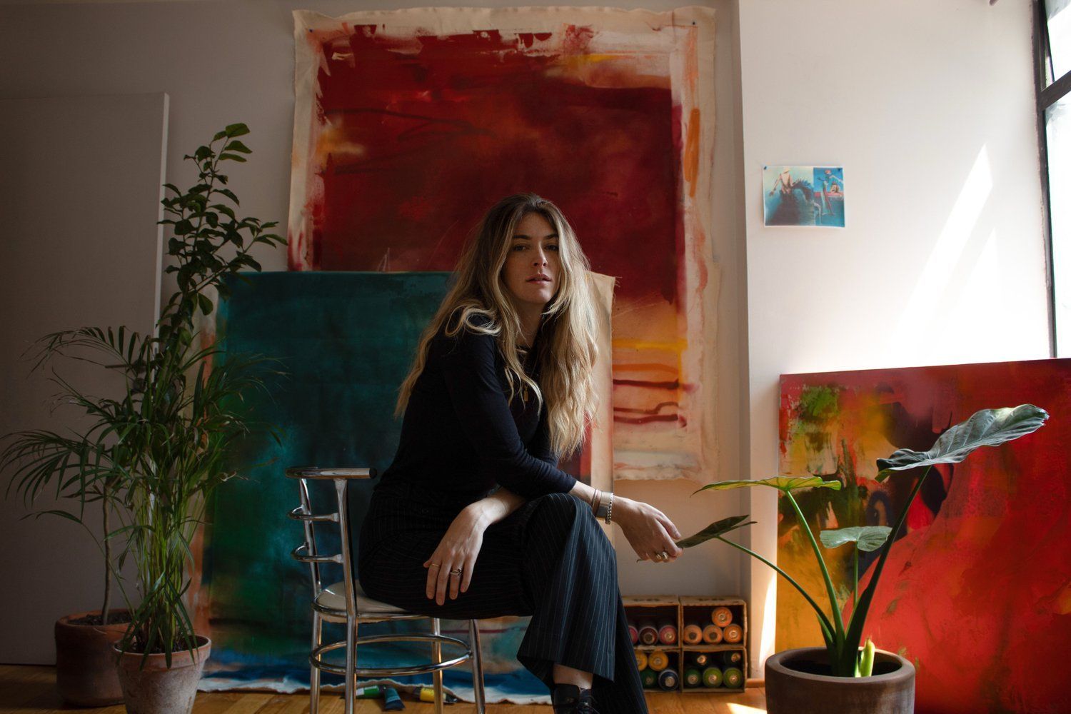 Woman seated in a studio with abstract red and teal paintings, plants, and soft natural light