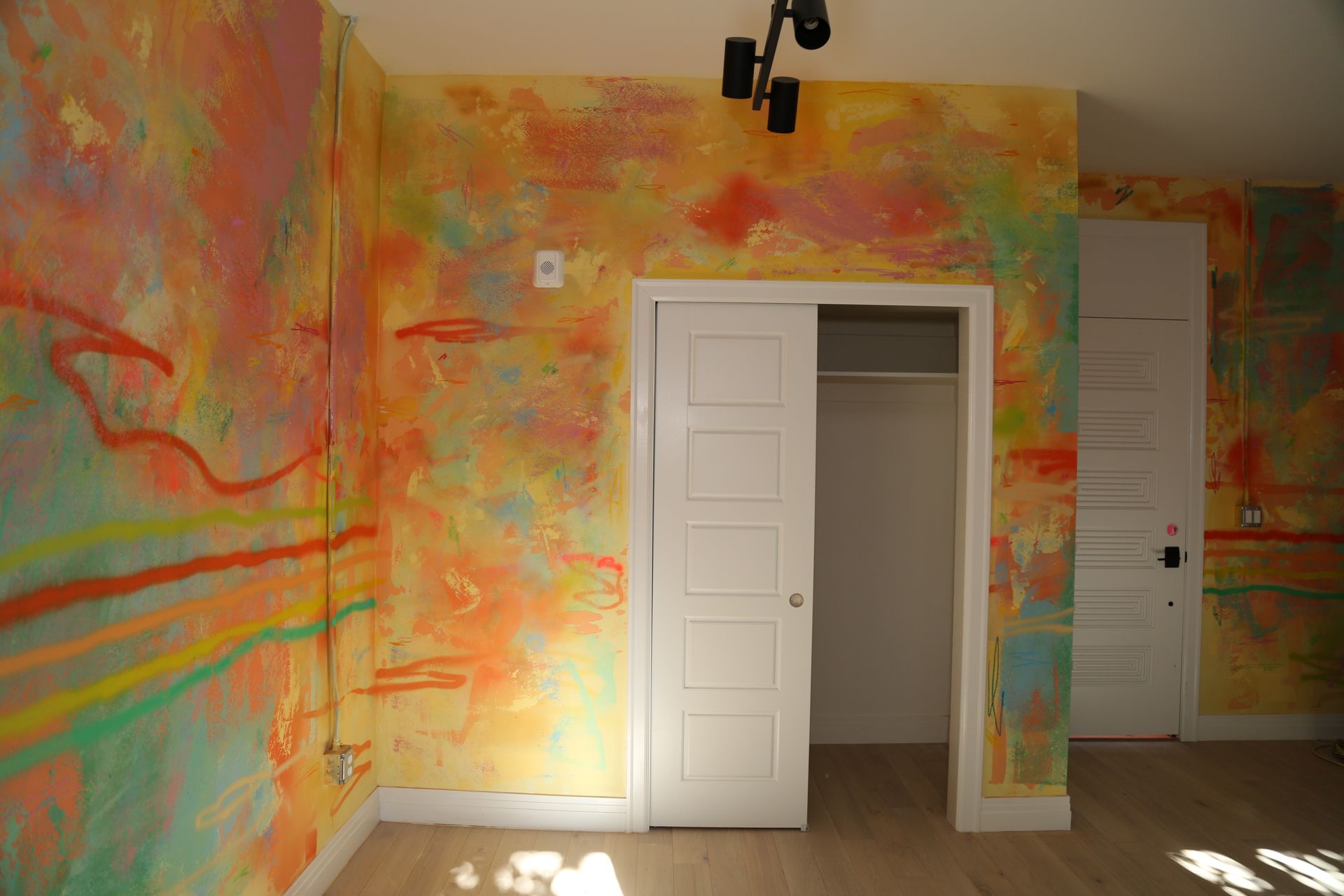 Empty room with colorful abstract paint on walls and white closet doors, lit by sunlight