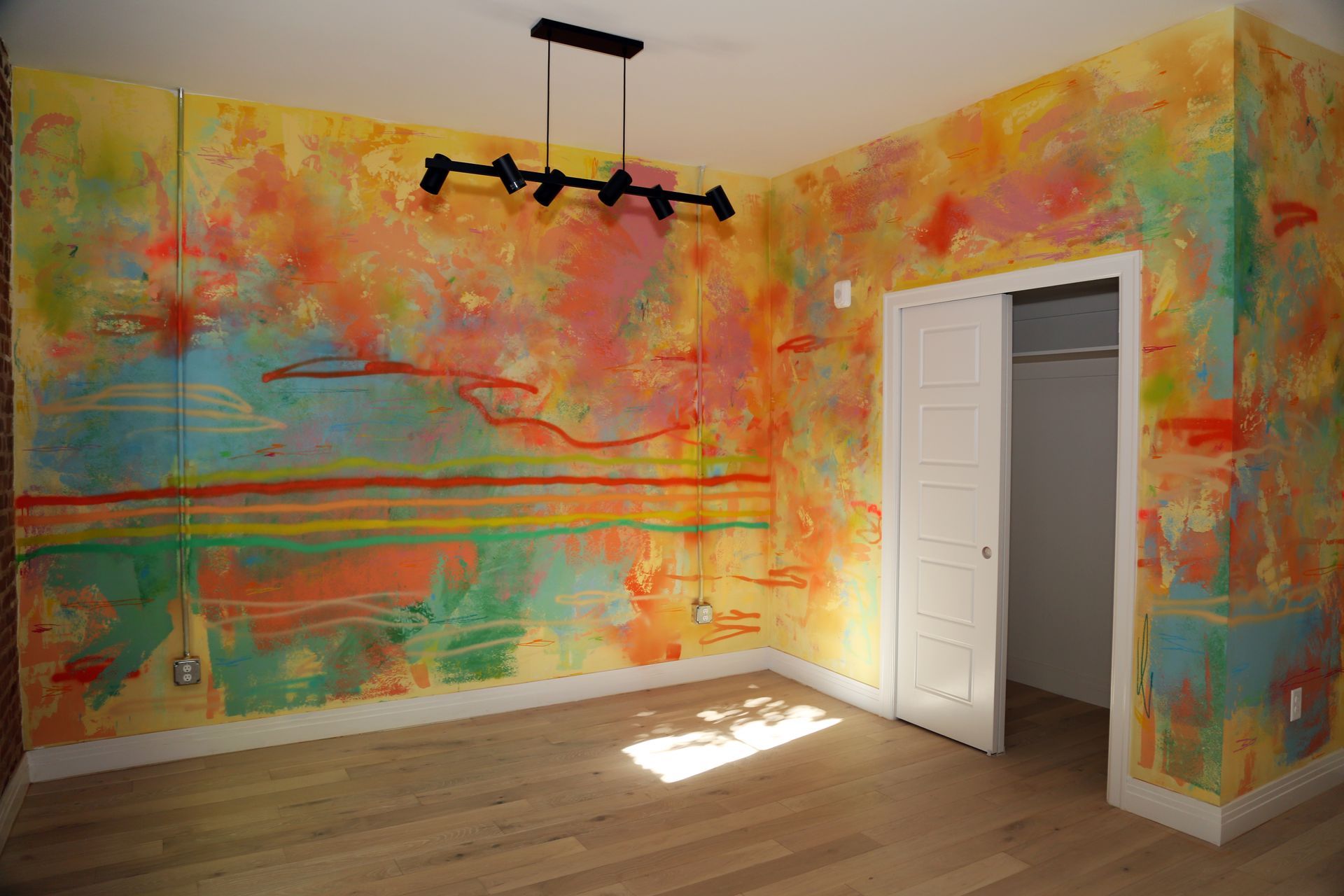 Colorful abstract mural in an empty room with wood floors, a white closet, and a modern ceiling light.