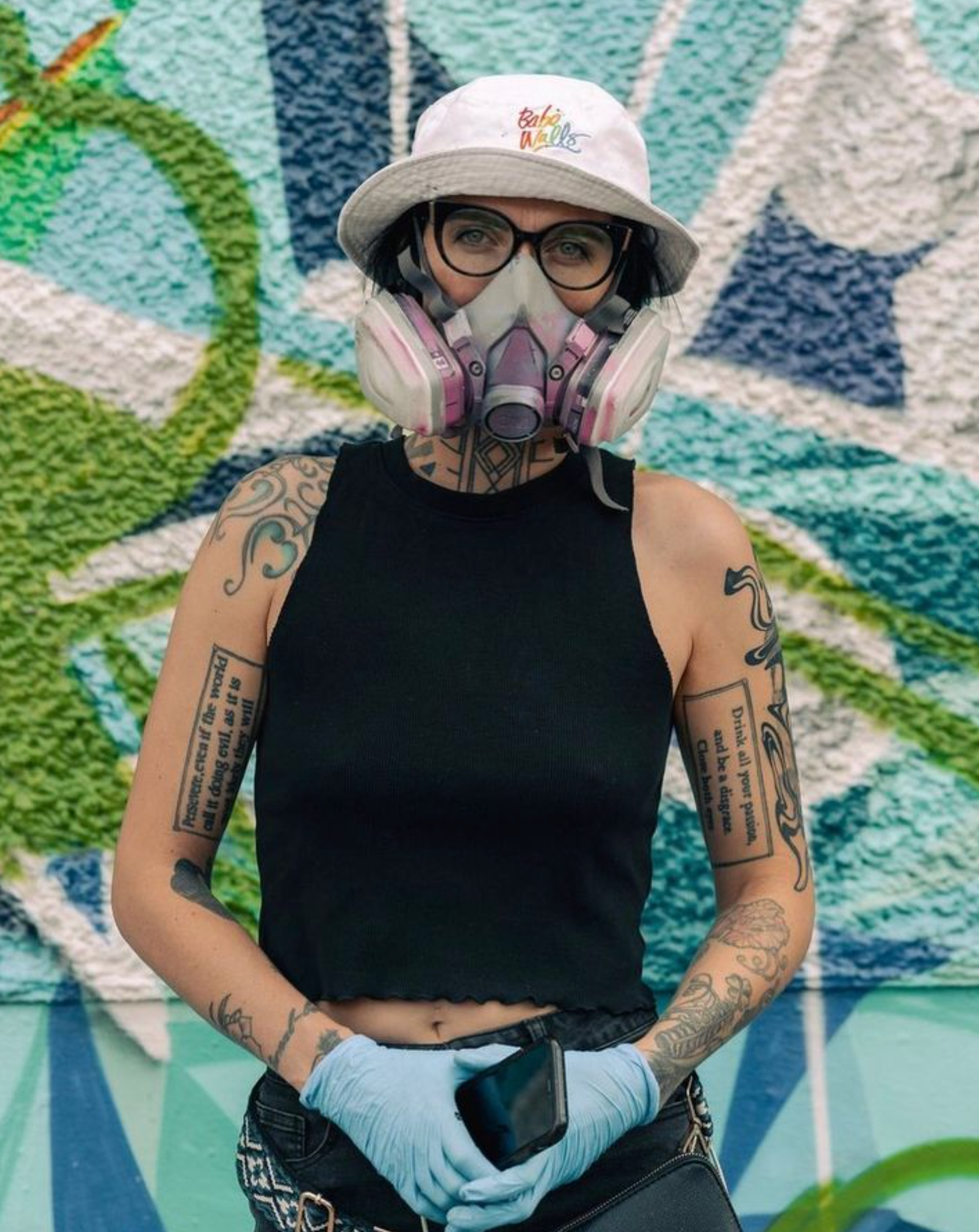 Person in hat, glasses, and respirator mask stands against a colorful graffiti wall.