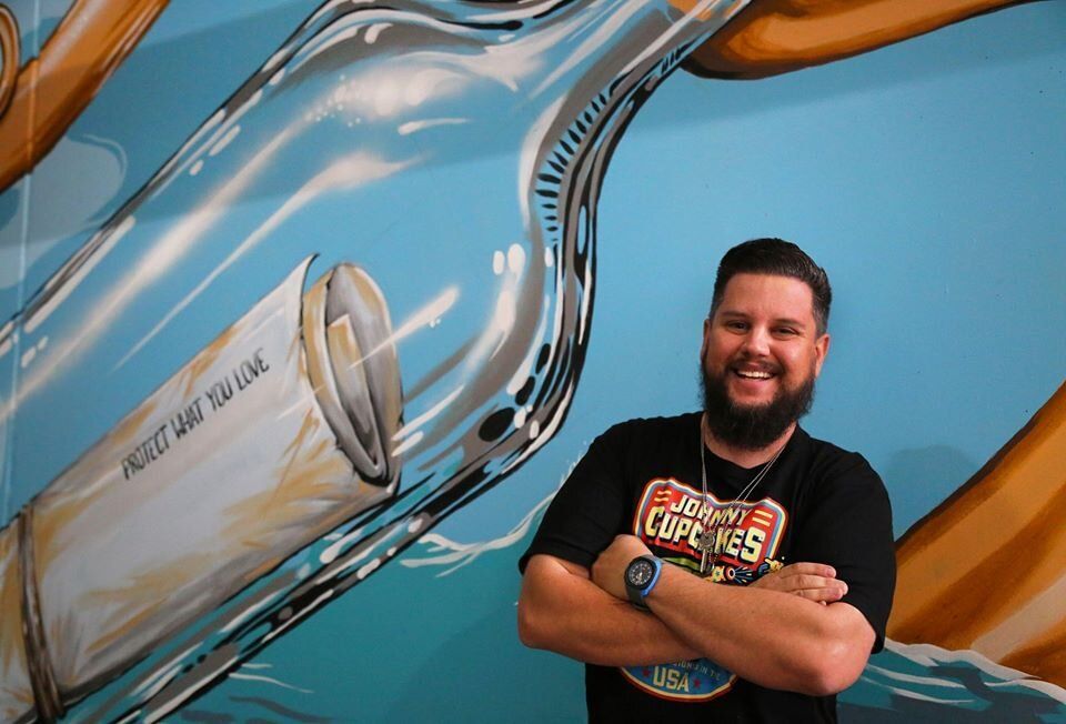 Smiling man with crossed arms posing before a blue mural of a giant bottle and glass