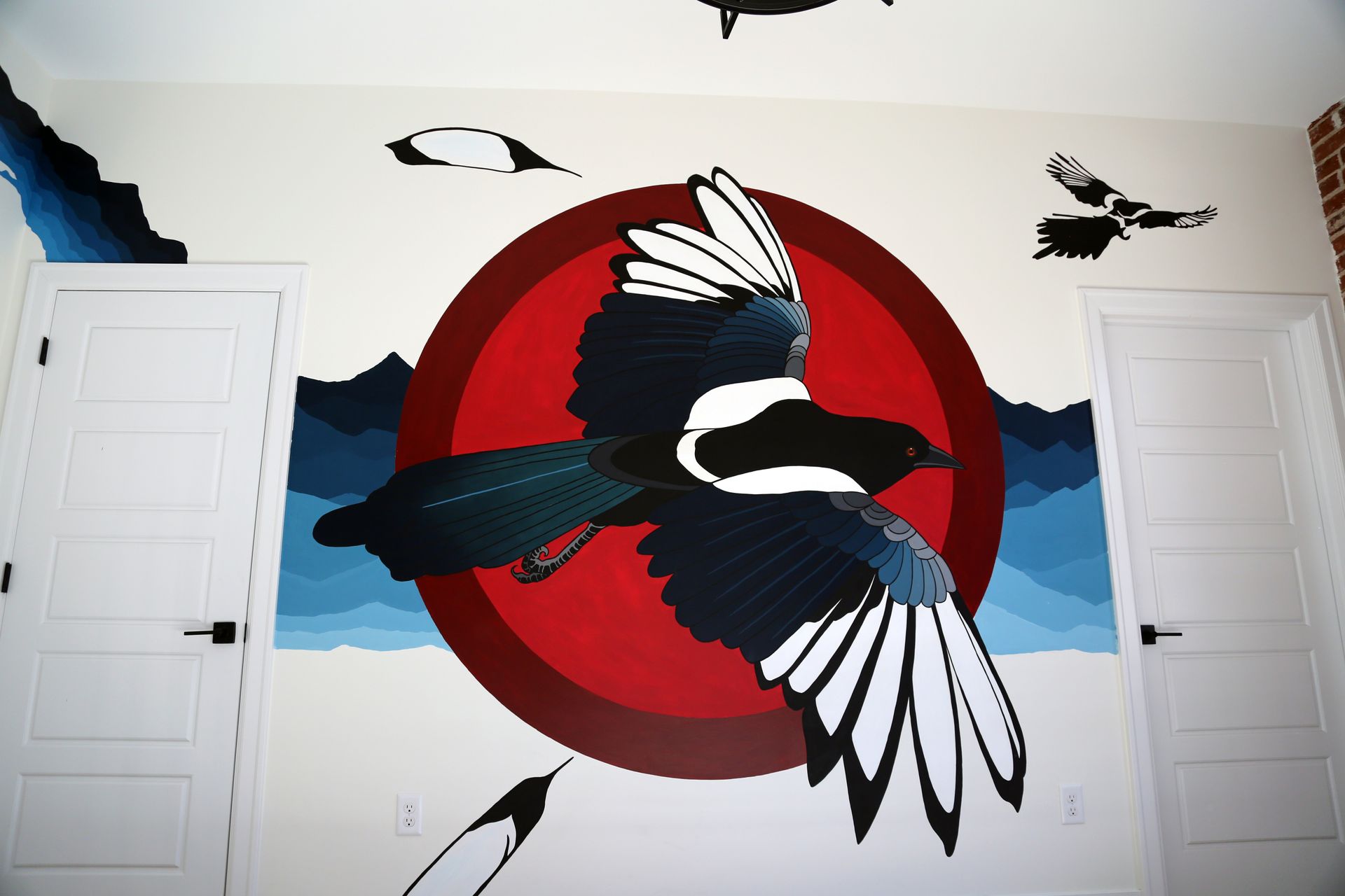 Mural of a black-and-white bird flying across a red sun on a white wall with doors