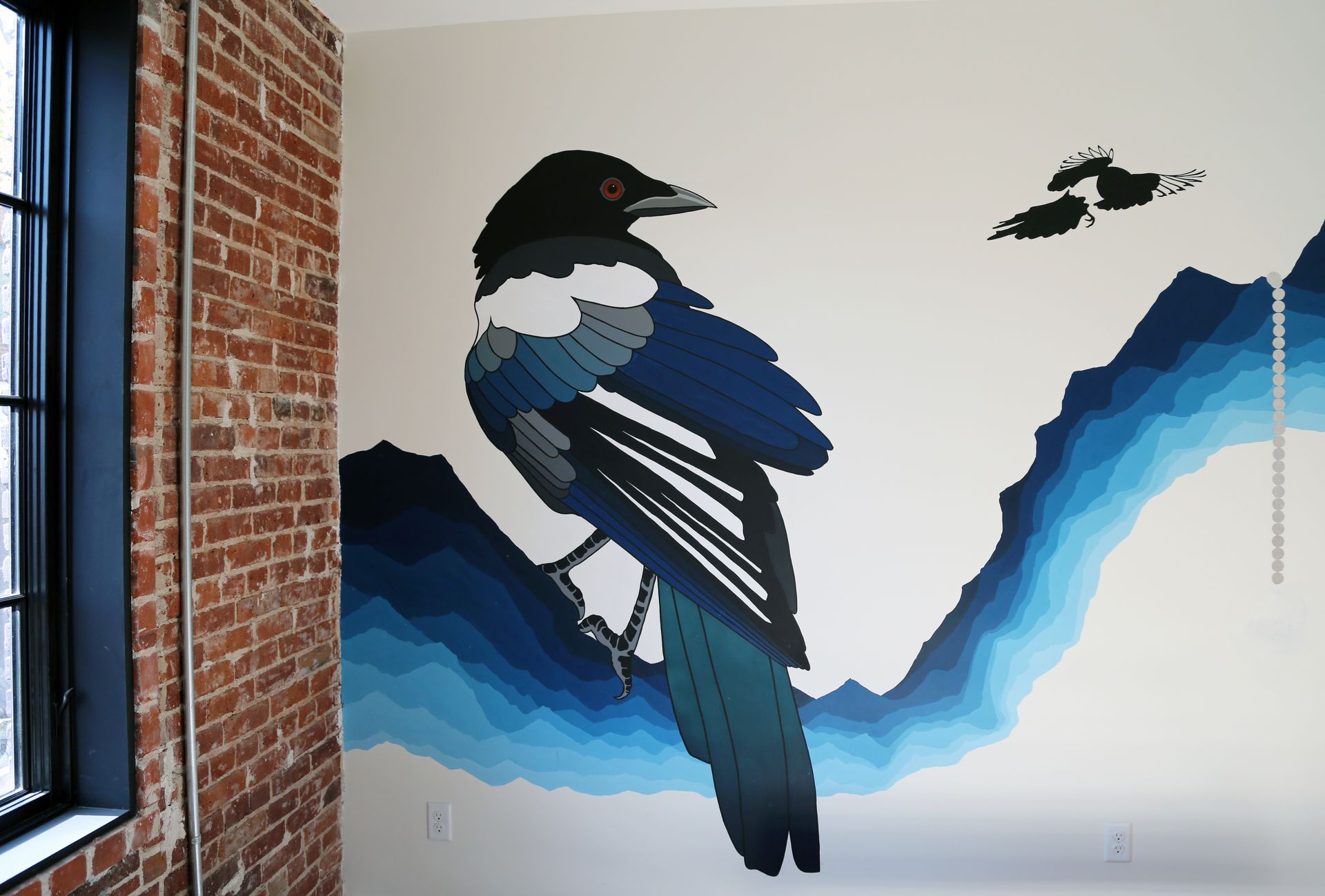 Mural of a black, white, and blue bird on a white wall beside a brick window frame